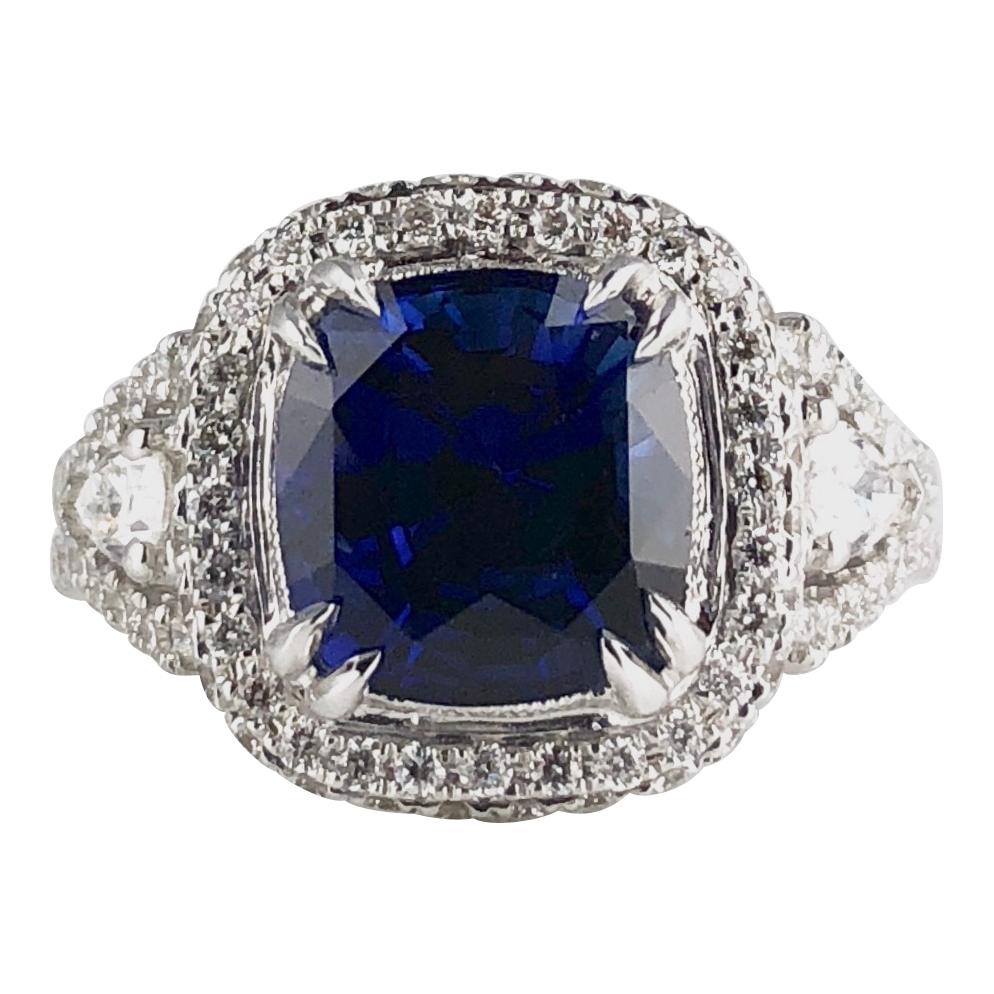 This ring sparkles with a 3.59 carat Cushion Cut Blue Sapphire center, surrounded by a halo of round white diamonds. The split shank starts with a pear shaped diamond, surrounded by round white diamonds, which also follow through the front and back