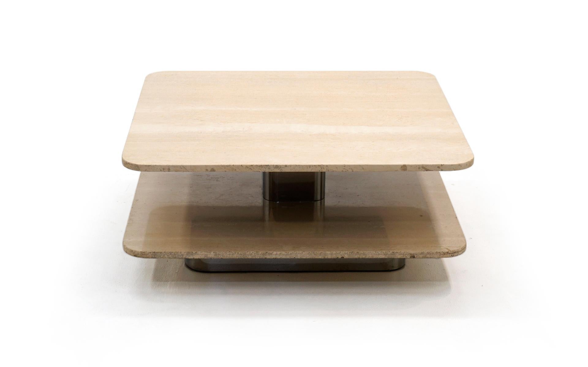 Table basse en travertin à deux niveaux, carrée et aux angles arrondis. La partie inférieure en travertin est fixée à une base chromée encastrée. Cette structure se poursuit au centre du travertin avec un support en acier chromé encore plus