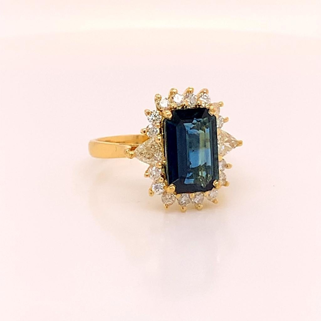 This timeless treasure is made of a phenomenal 3.65 Carat Emerald Cut Teal Sapphire surrounded by a unique halo of glittering diamonds made up of both Round Brilliant and Trillion Cut diamonds weighing 0.85 Carats in total. The extraordinary beauty