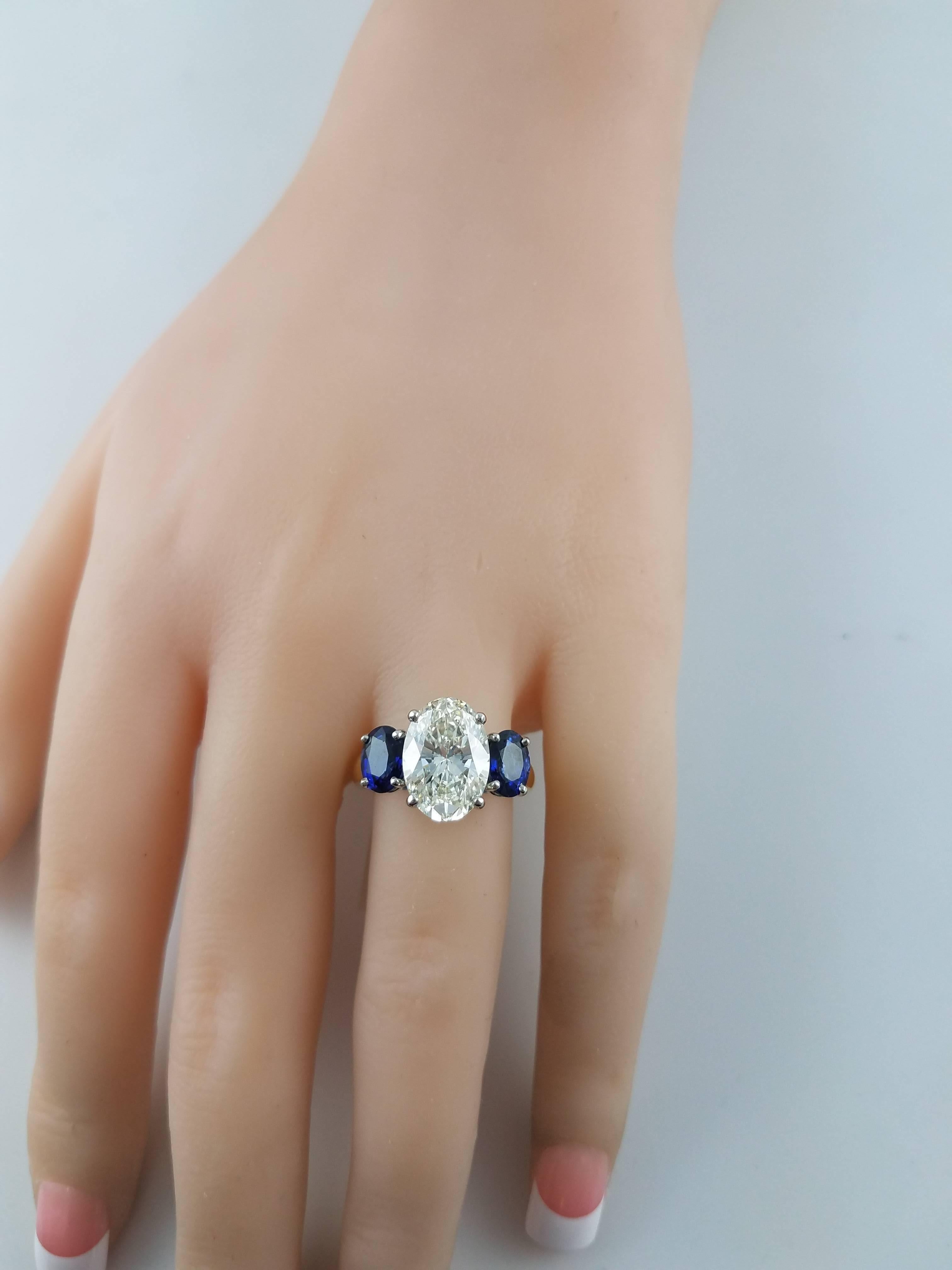 An engagement ring like no other. Showcasing a sparkling oval cut diamond center stone weighing 3.69 carats. Flanking the center are two vibrant blue sapphires each set in a platinum basket. Weight of the sapphires 2.08 carats total. The rounded