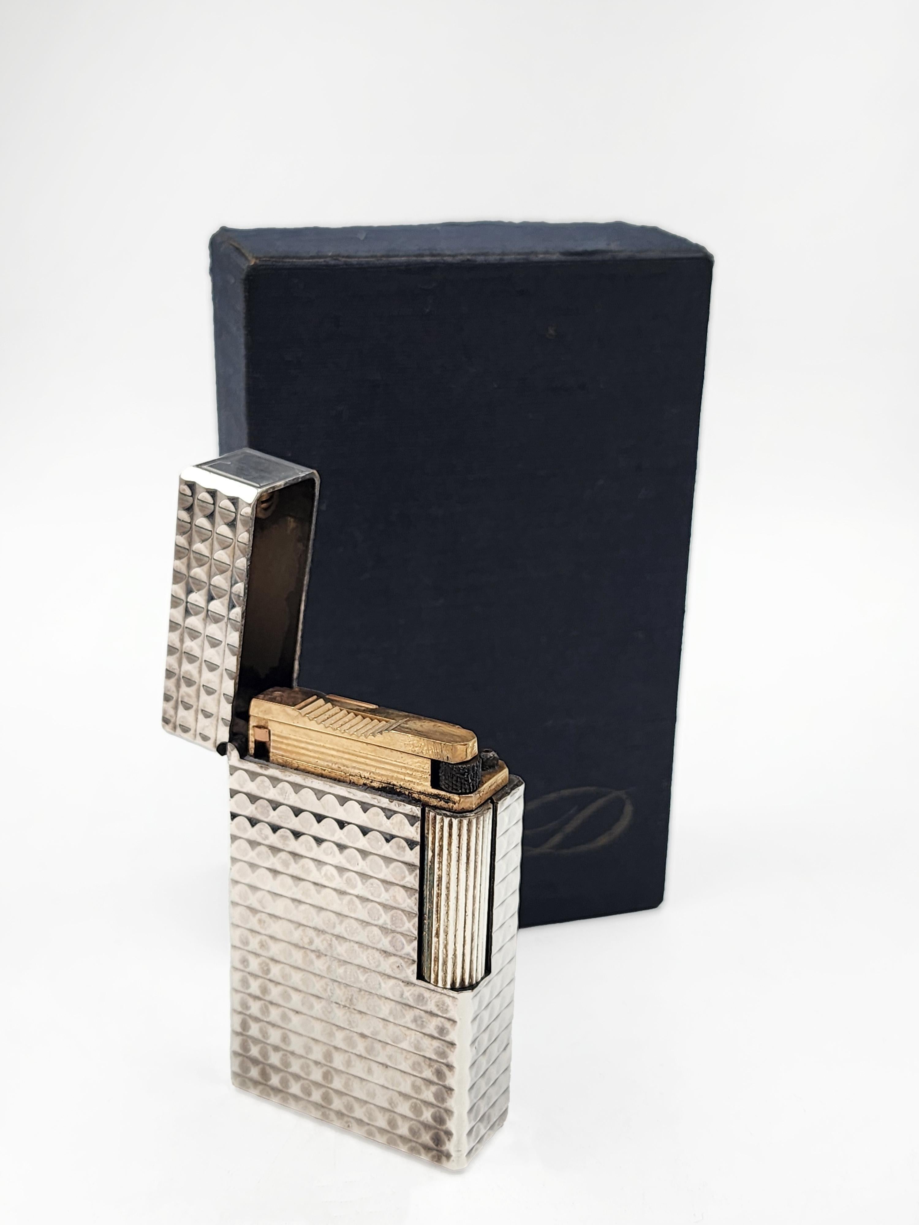 This is a vintage 1970s S.T. Dupont lighter, made in France, with a silver finish and a textured geometric pattern on the casing. It comes with its original blue velvet pouch and a black outer box. The base of the lighter displays the 