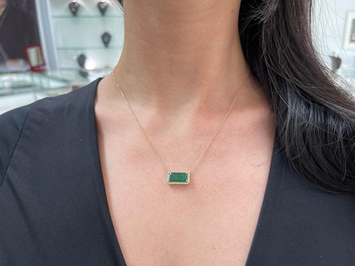 Displayed is a stunning emerald and diamond halo pendant totaling 3.91-carats. Handcrafted in 14k yellow gold, this pendant features a dark rich green, elongated emerald cut-emerald. Surrounding the rare earth-mined gemstone are natural, brilliant