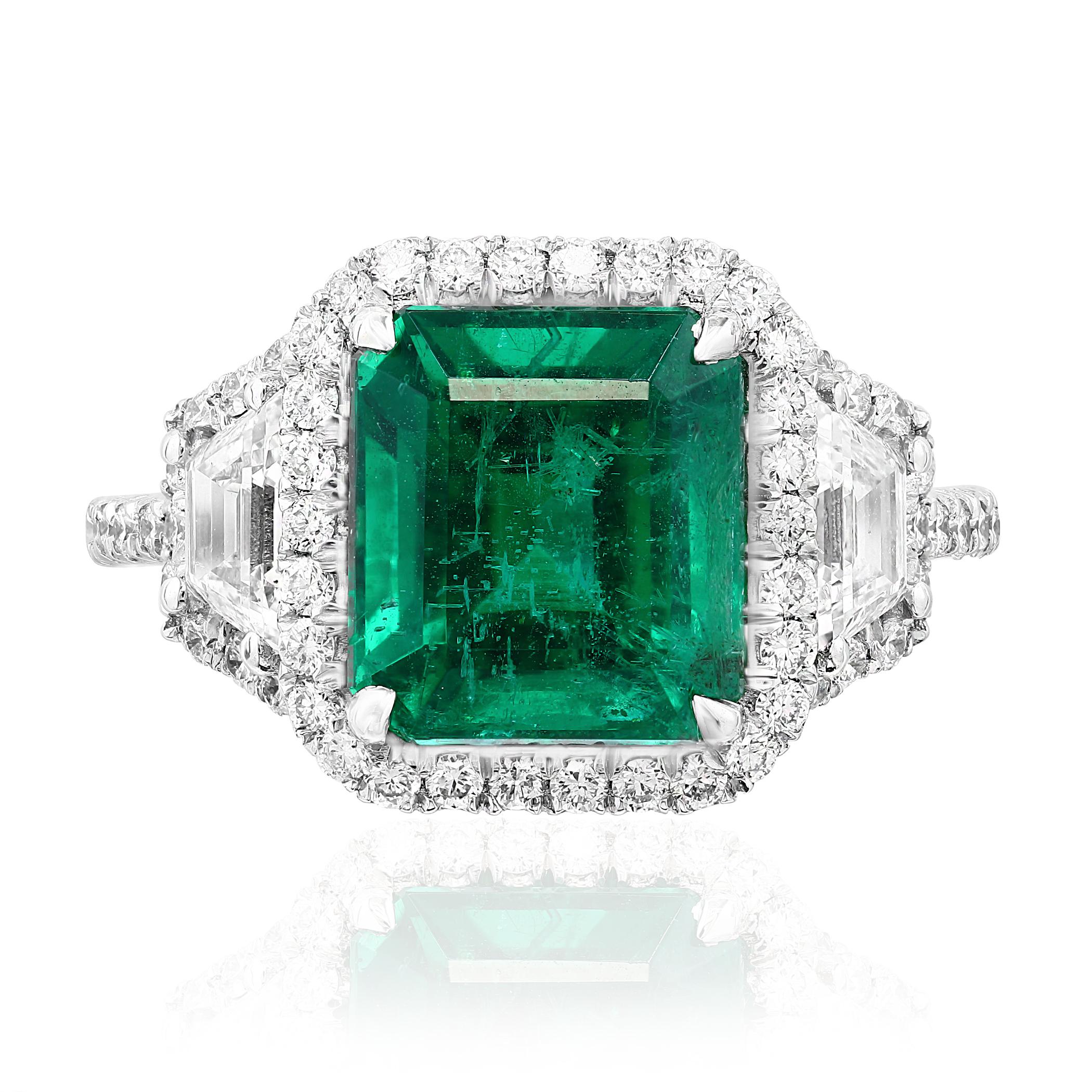 A stunning ring showcasing a rich green emerald cut emerald weighing 3.93 carats surrounded by a row of diamonds. Flanking the center stone are two brilliant cut trapezoid diamonds, weighing 0.71 carats total, framed in a brilliant diamond halo. 58