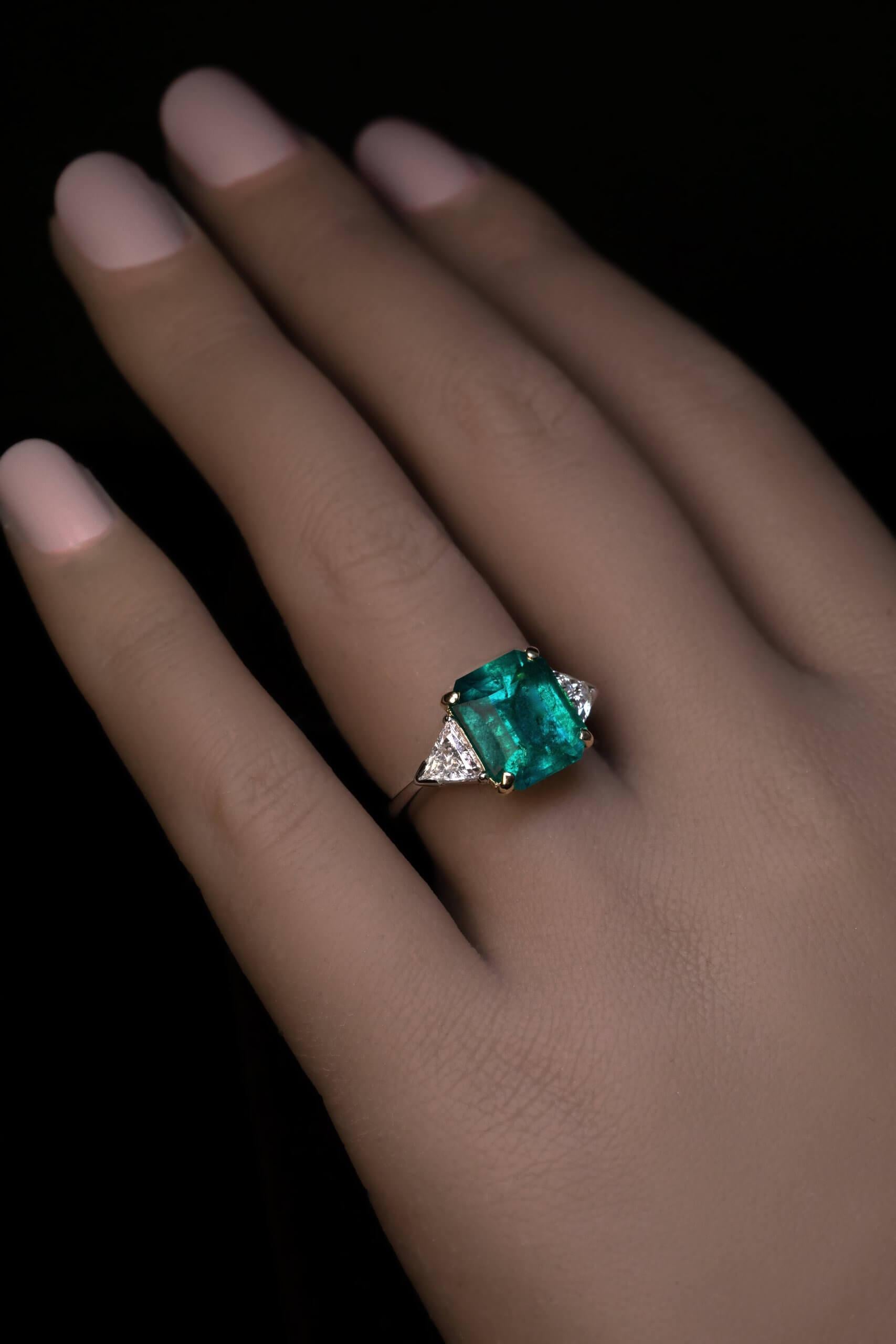 This contemporary platinum and gold ring features a 3.96 carat Colombian emerald of a vivid bluish green color flanked by two trillion cut diamonds. The emerald has good color saturation and sparkle.  The actual color of the emerald is more vivid