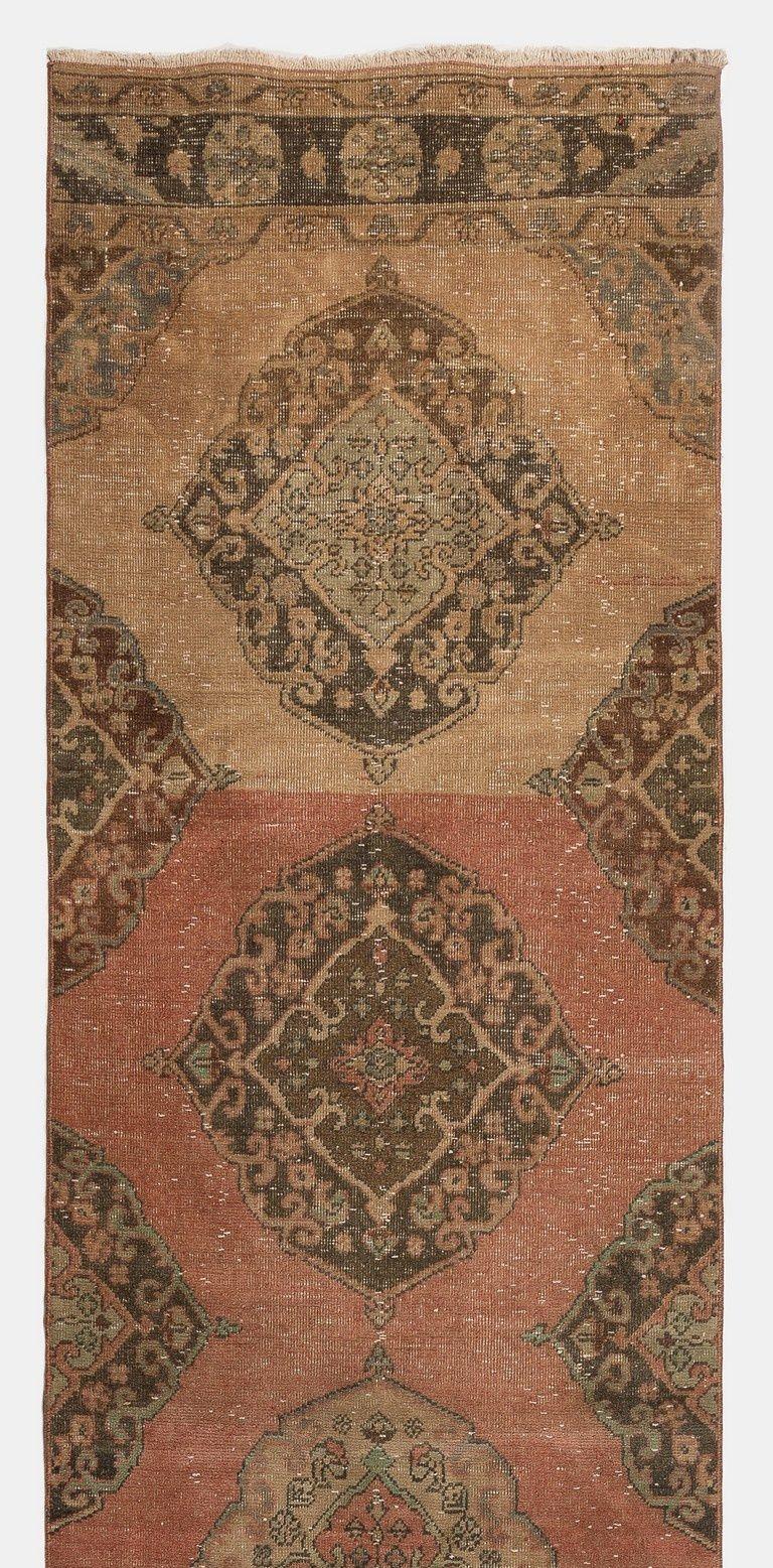 This vintage Turkish runner rug was hand-knotted in the 1960s. It features multiple full and half medallions decorated with floral motifs in brown, peach and sage green against a plain background in peach and dark coral. It has medium wool pile on