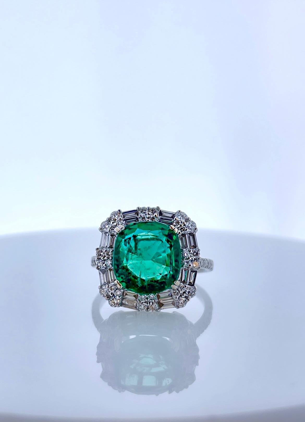 This is a 4.06CT cushion cut intense green emerald. The gem source is Colombia; its color is vibrant grass green. Its transparency and luster are excellent. Sparkling mixed cut diamonds frame this gem in a halo cluster and down the sides of the slim