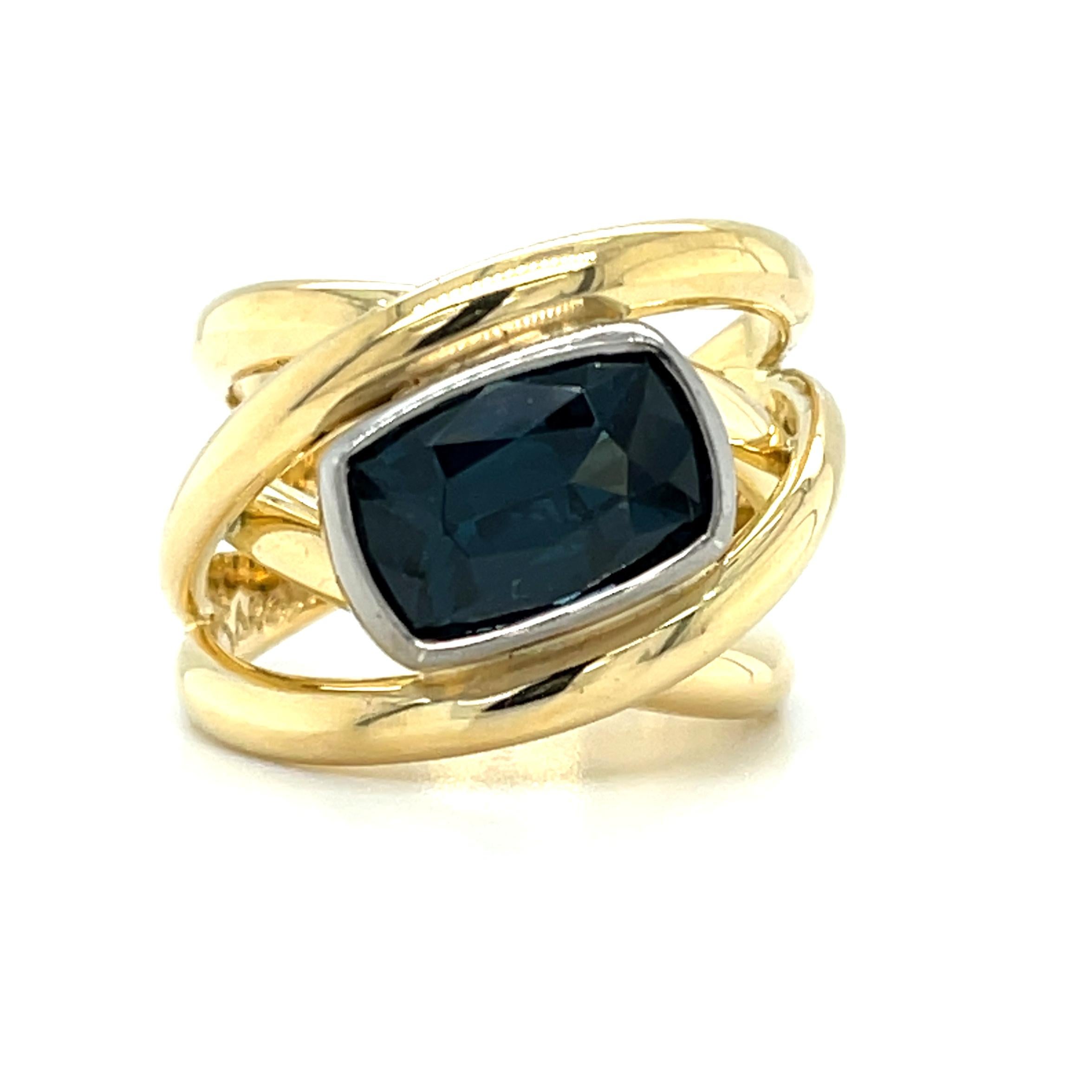 This striking band ring features a 4.07 carat cushion-shaped blue spinel with beautiful rich, navy blue color! The lovely gem is set on a horizontal diagonal, in an 18k white gold bezel. High-polished 18k yellow gold bands swirl around the center