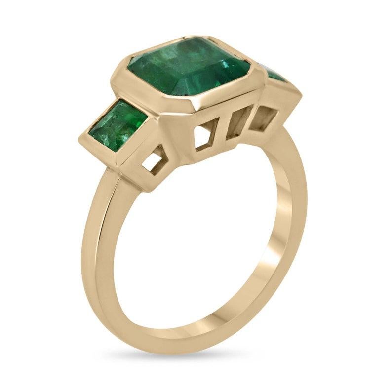 Introducing our exquisite Emerald Trilogy Ring, featuring a magnificent 3.08-carat Asscher-cut emerald as the focal point, radiating with a rich dark green hue and transparent clarity. Complemented by a princess-cut emerald on each side, set in