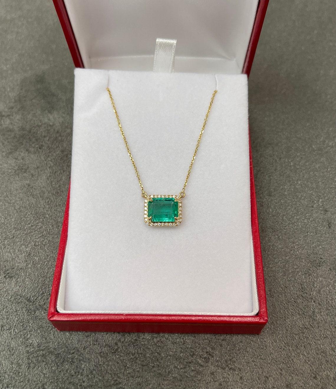 Featured here is a stunning statement size East to West rich bluish green emerald and round diamond halo necklace handmade in fine 14K yellow gold. Displayed is a lustrous, medium rich green with very good transparency, accented by a simple secure