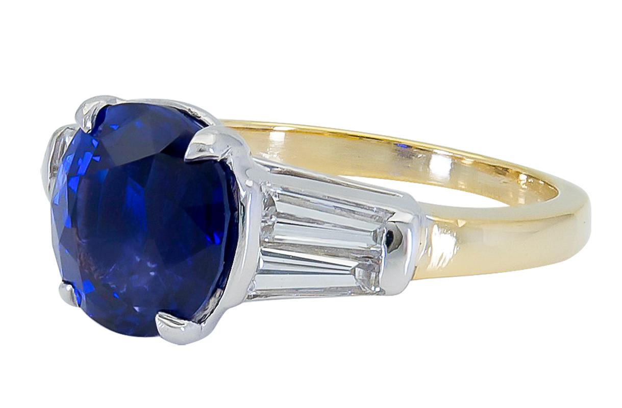 Ein zeitloser Verlobungsring mit einem runden blauen Saphir im Kissenschliff, flankiert von zwei spitz zulaufenden Baguette-Diamanten auf beiden Seiten. Eingefasst in eine Fassung aus 18 Karat Gold und Platin. 
Der blaue Saphir wiegt 4.11 Karat.
Die