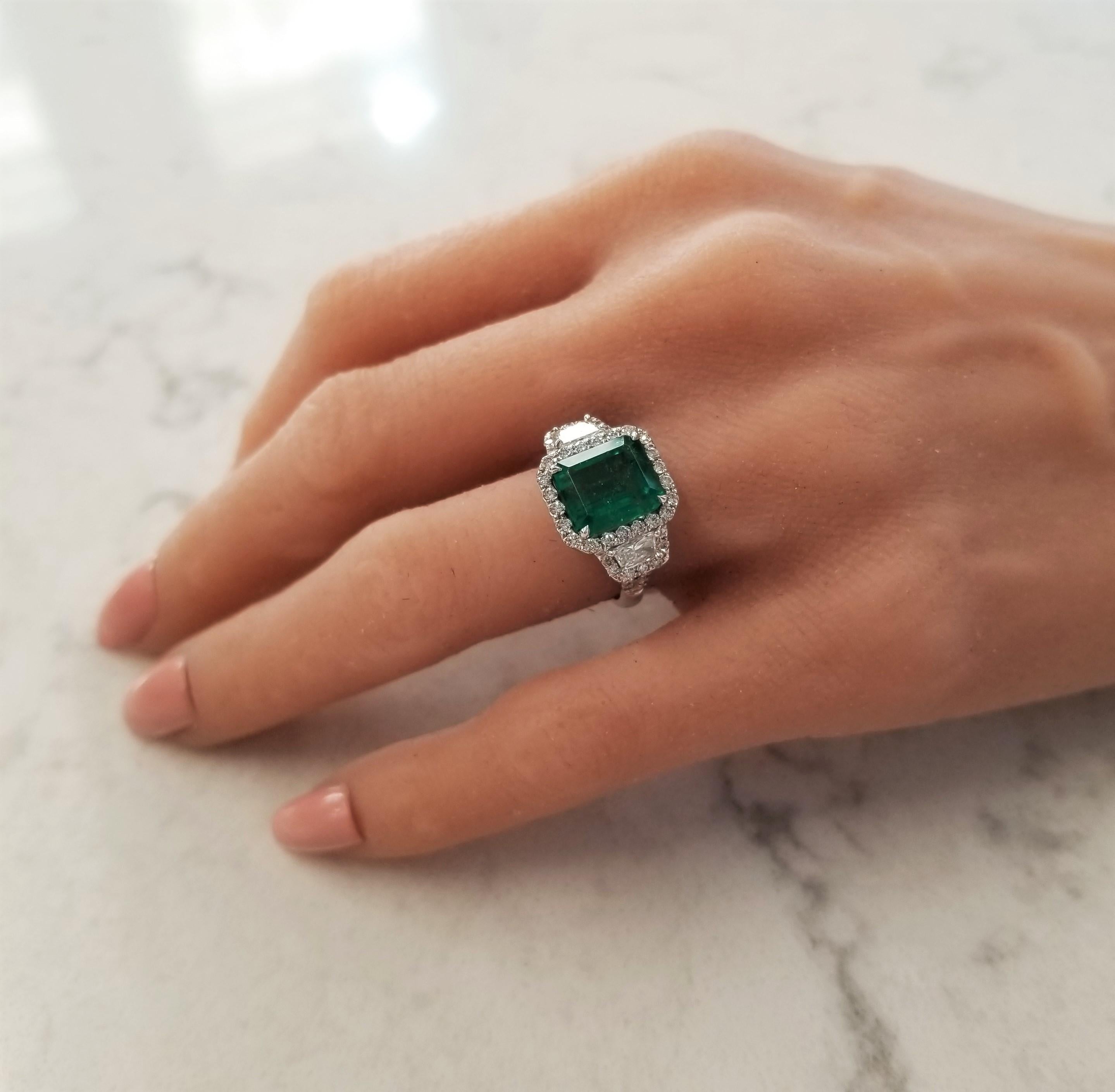 This beautiful 4.11 carat, emerald step-cut emerald is the spotlight. This emerald is from Brazil; its transparency is excellent, color is vibrant. This stellar three-stone ring is paired with 0.48 carats of trapezoid diamonds and glittering with an