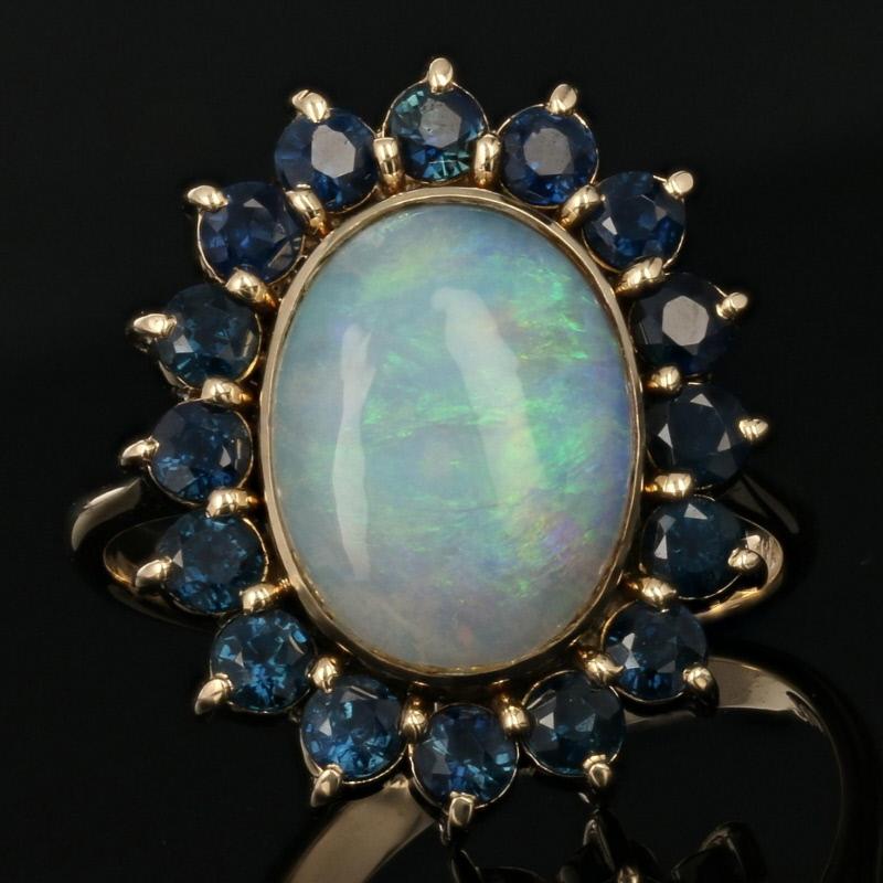 This ring’s captivating beauty will continue to dazzle long after sunset! Featuring a glamorous halo design, this 18k yellow gold piece showcases a mesmerizing opal cabochon framed by sumptuous blue sapphires. 

This ring is a size 7 3/4, but it can