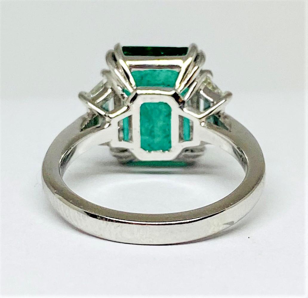 Beautiful three stone engagement ring in 18KT white gold featuring an incredibly gorgeous 4.18 carat GIA certified step cut prong set deep green emerald center set between two trapezoid shaped white diamonds weighing a total of 1.20 carats on an