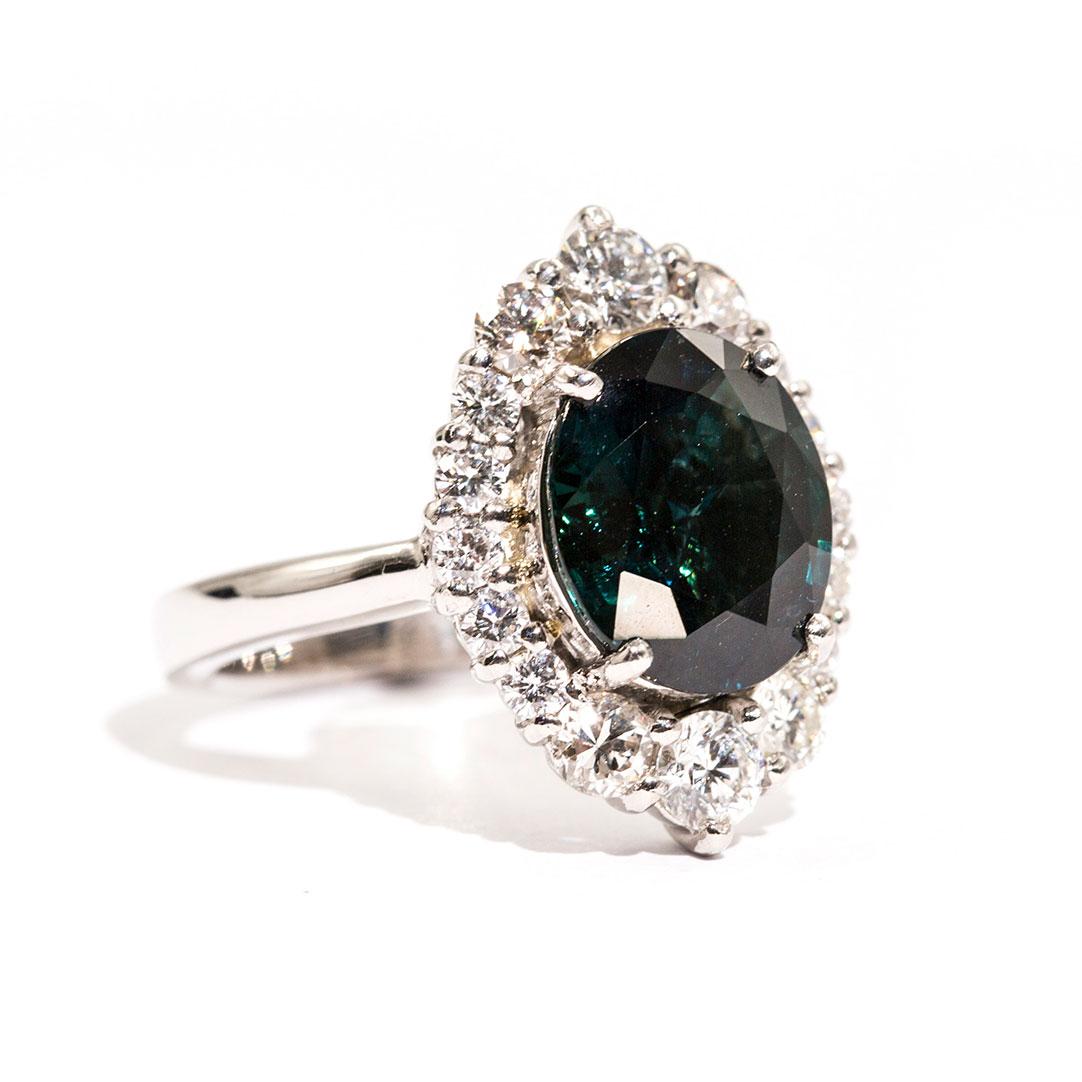 Forged in platinum is this wondrous ring that features a breath-taking 4.25 carat oval natural sapphire of a strong deep teal (blue green) colour complimented with an alluring border of total of 1.16 carats of sparkling round brilliant cut diamonds.