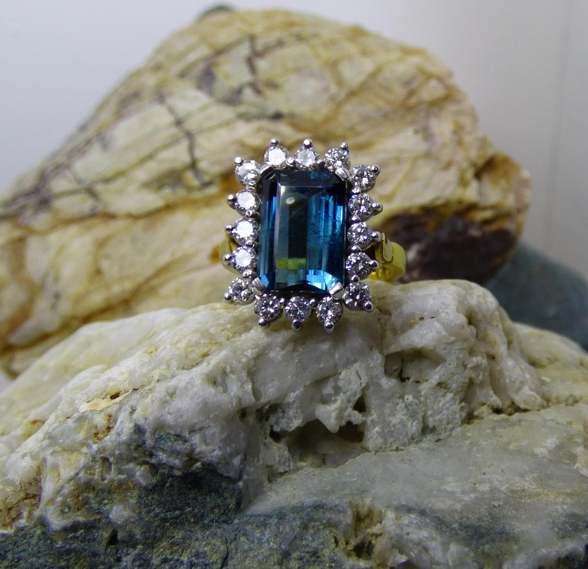 A clean bright and colourful Indicolite (Blue Tourmaline) is 12X9mm ( 4.25ct.)is emerald cut. The Tourmaline is surrounded by 16 Diamonds with a total weight of .81ct.  The ring is handmade in 18K yellow gold with the Diamonds set in white gold. 