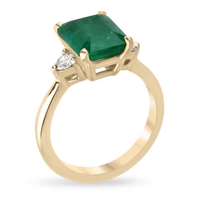 This exquisite three-stone ring showcases a stunning emerald-cut emerald set north to south in four prongs, radiating a rich vivid green hue with semi-transparent clarity. Flanked by petite brilliant round-cut diamonds on each side, the emerald’s