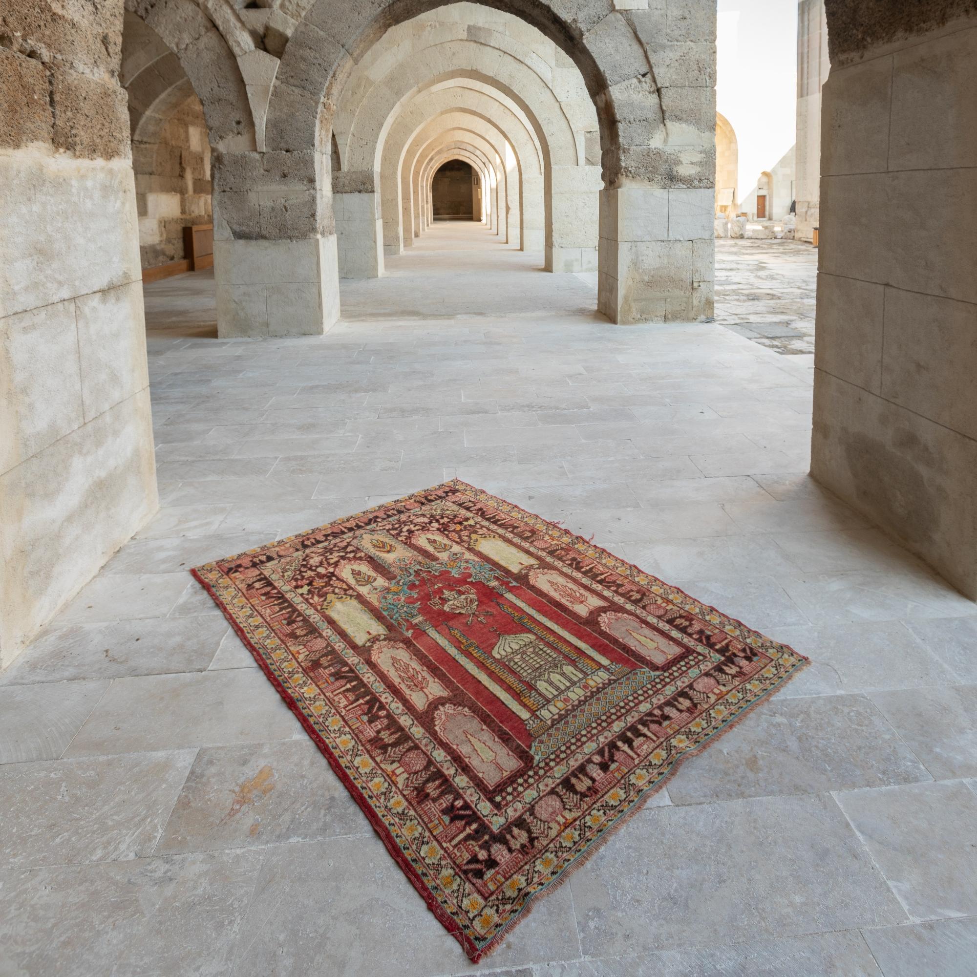 A one-of-a-kind semi antique Anatolian prayer rug featuring a carefully constructed arch design, framing at its base the depiction of a mosque that stands under a large pendant lamp and between two grand columns reaching into the ornate crown of the