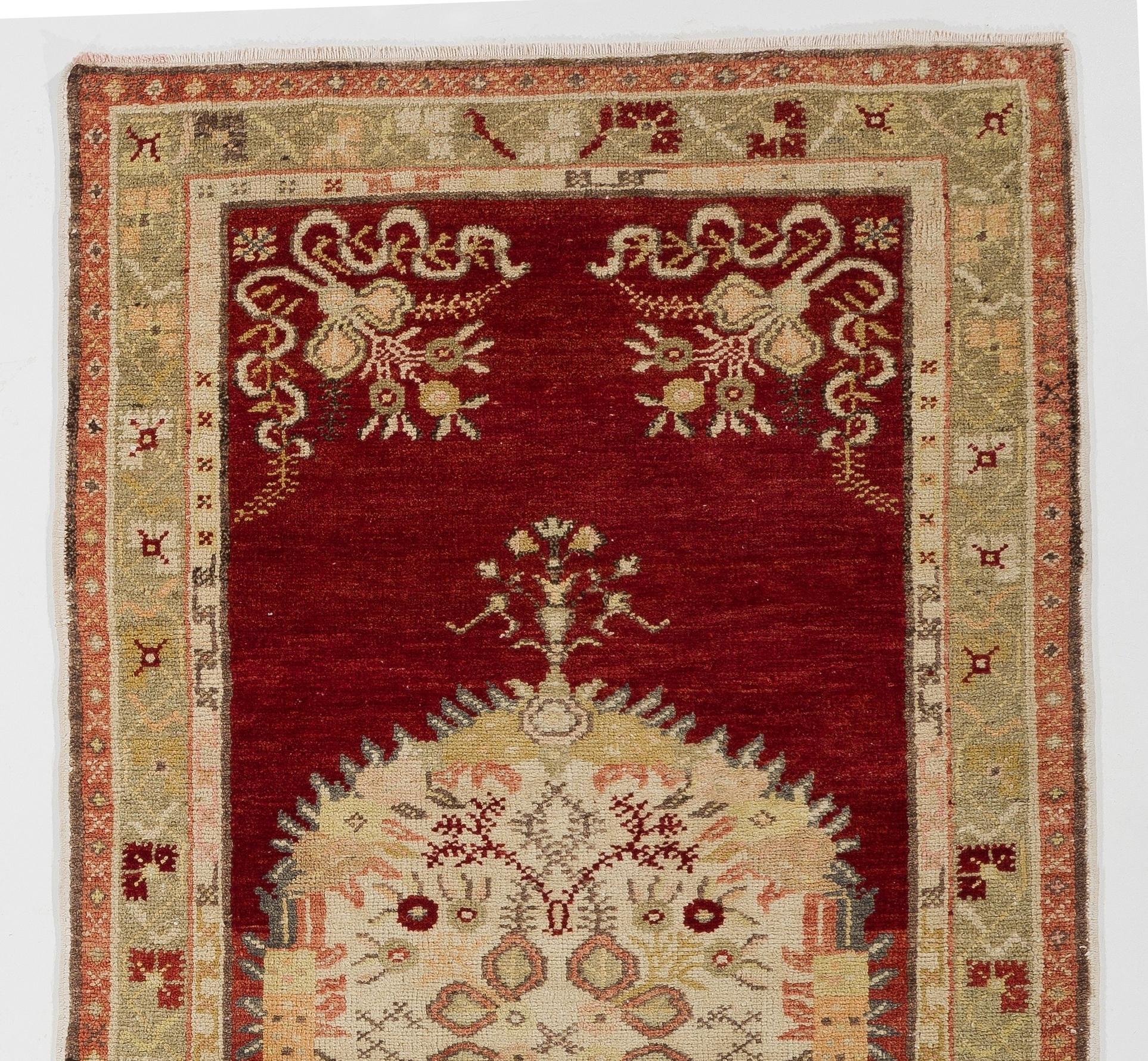 This vintage hand knotted Turkish accent rug boasts a large elliptical medallion at its centre in ivory, pale yellow, light olive green, soft orange and salmon pink. It is decorated with stylized florals against a deep, ruby red field that features