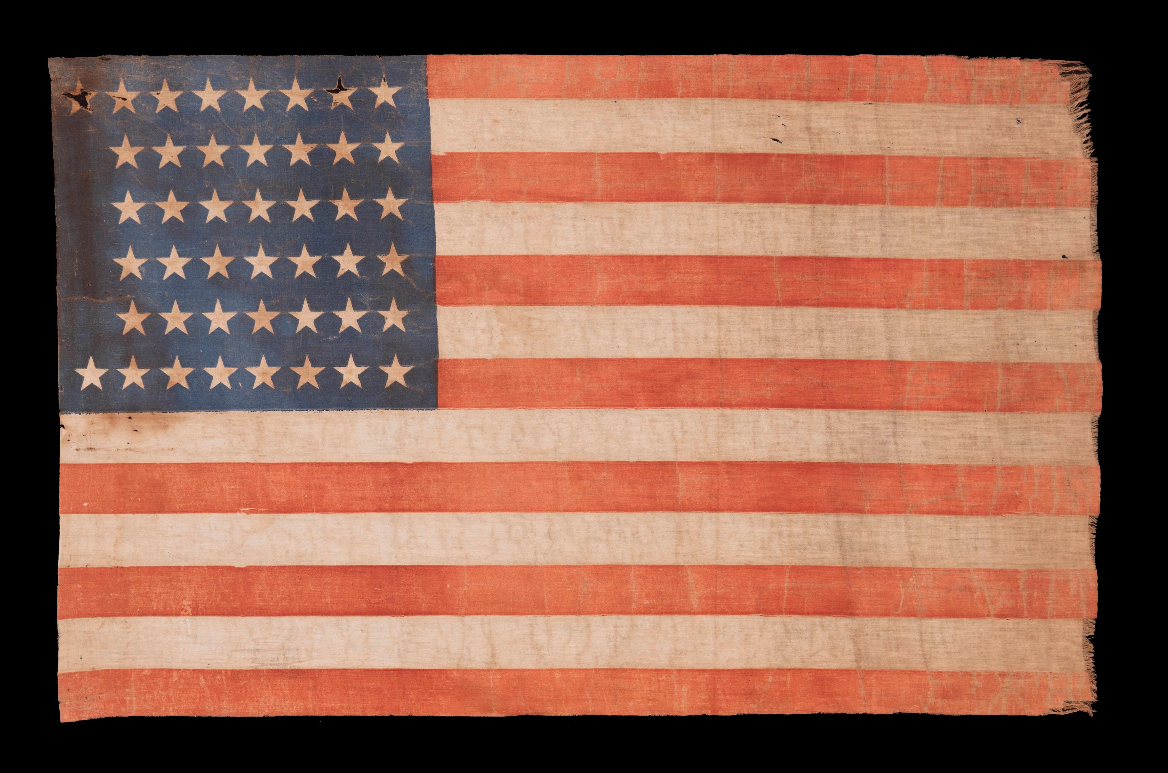 44 STAR ANTIQUE AMERICAN PARADE FLAG WITH ENDEARING WEAR FROM LONG TERM USE AND WITH ITS STARS ARRANGED IN A NOTCHED PATTERN THAT LEAVES 4 SPACES OPEN FOR THE REMAINING WESTERN TERRITORIES, REFLECTS THE PERIOD WHEN WYOMING WAS THE MOST RECENT STATE