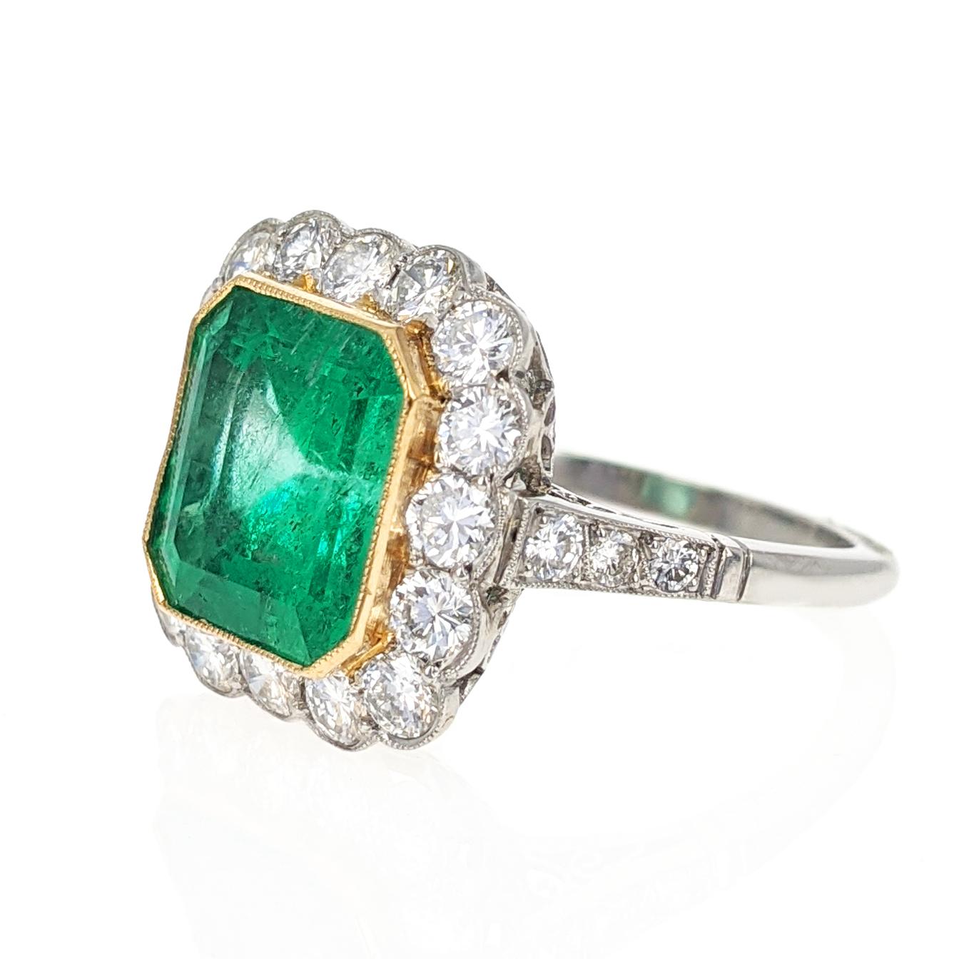 This beautiful ring centers upon a 4.43 carat octagonal step-cut emerald, bezel set in yellow gold, surrounded by 16 round brilliant-cut diamonds, further flanked by 3 round brilliant-cut diamonds on each of the shoulders. Total diamond weight is