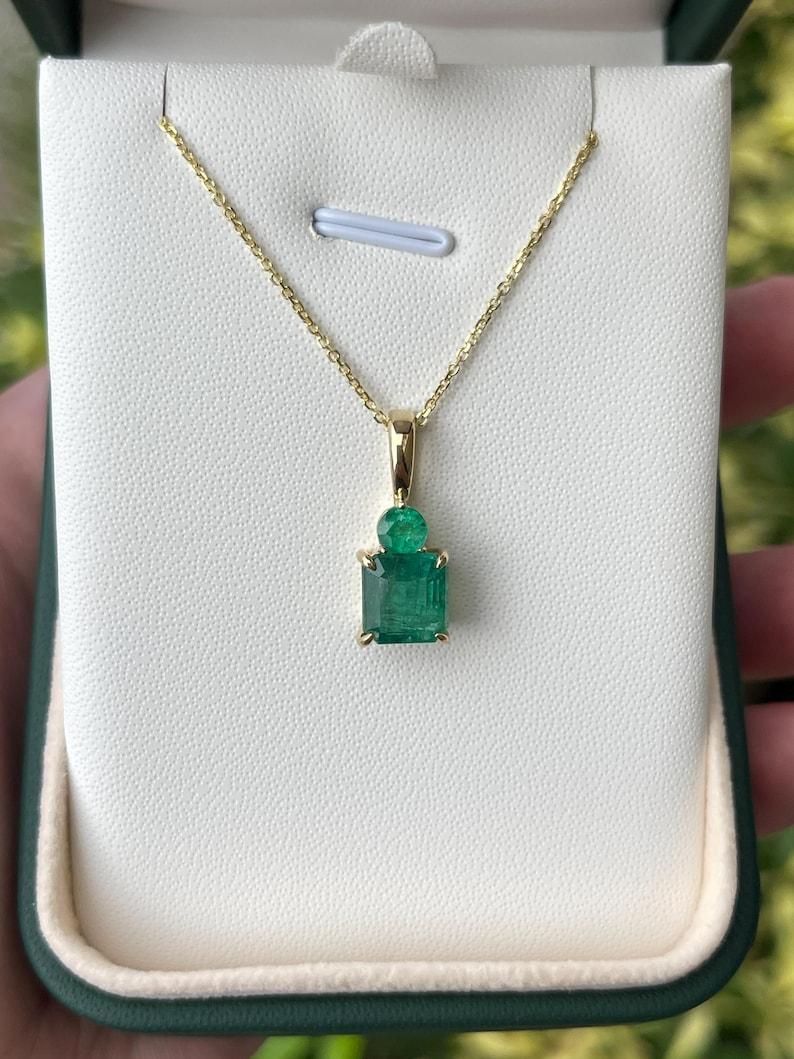 Elevate your neckline with the captivating beauty of our emerald Toi et Moi pendant. Showcasing two stunning natural Zambian emeralds totaling 4.86 carats, this pendant features an asscher cut emerald as its focal point, complemented by a sizable
