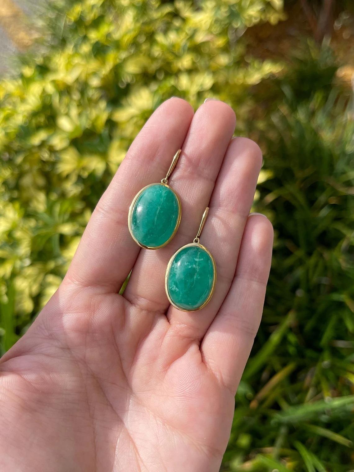 An impressive ONE OF A KIND set of solitaire huge Colombian emerald hook earrings in fine 18K yellow gold. Displayed are ethically earth-mined emeralds with rich deep green color accented by a lightweight, skillfully hand-made golden bezel. The 18K