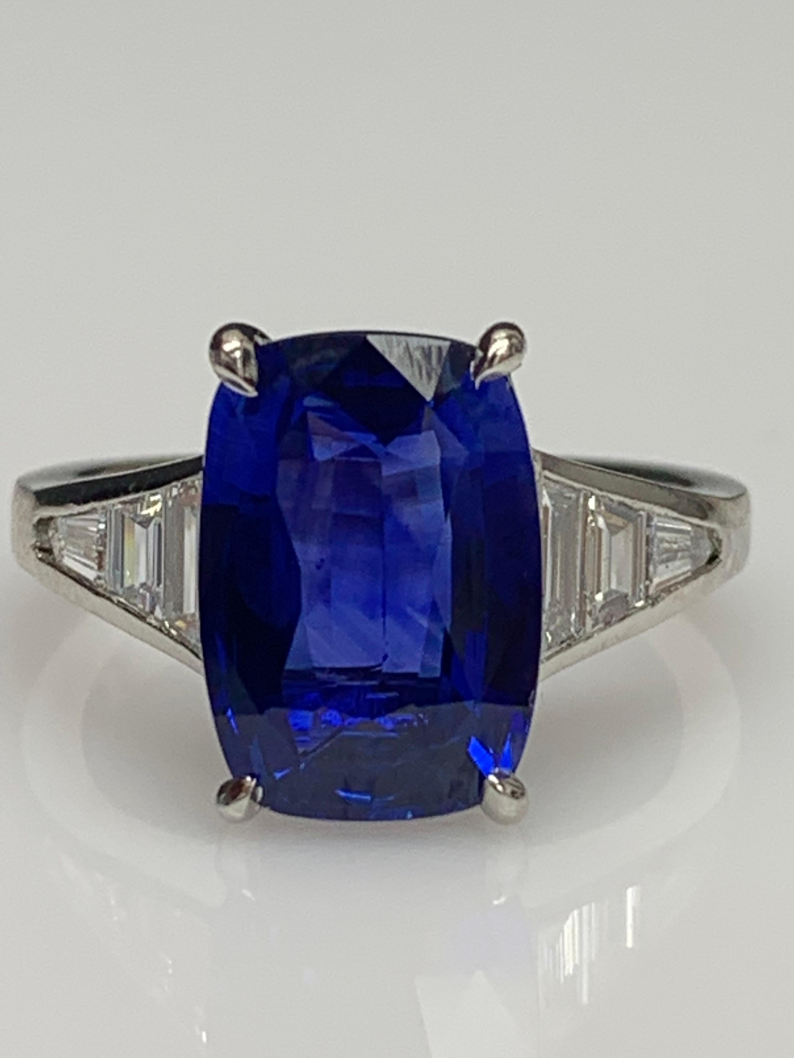 A stunning well-crafted engagement ring showcasing a 4.50 Carat elongated Cushion Blue Sapphire. Flanking the center stone are Perfectly matched graduating step-cut diamonds, channel set in a polished platinum mounting. 6 Accent diamonds weighing