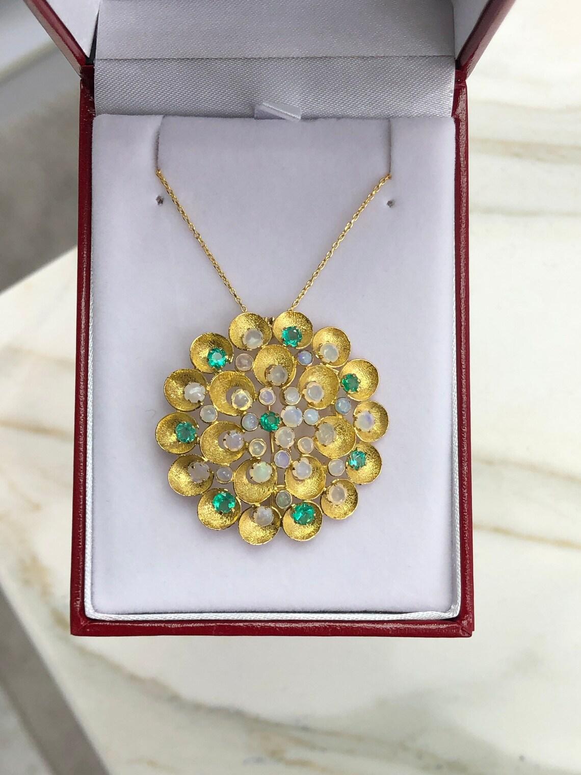 This pendant is not for the faint of heart! Featured here is a stunning Colombian emerald and opal pin/pendant made in fine 14K yellow gold. Displayed are lustrous, medium-light green emeralds with incredible transparency, accented by a simple