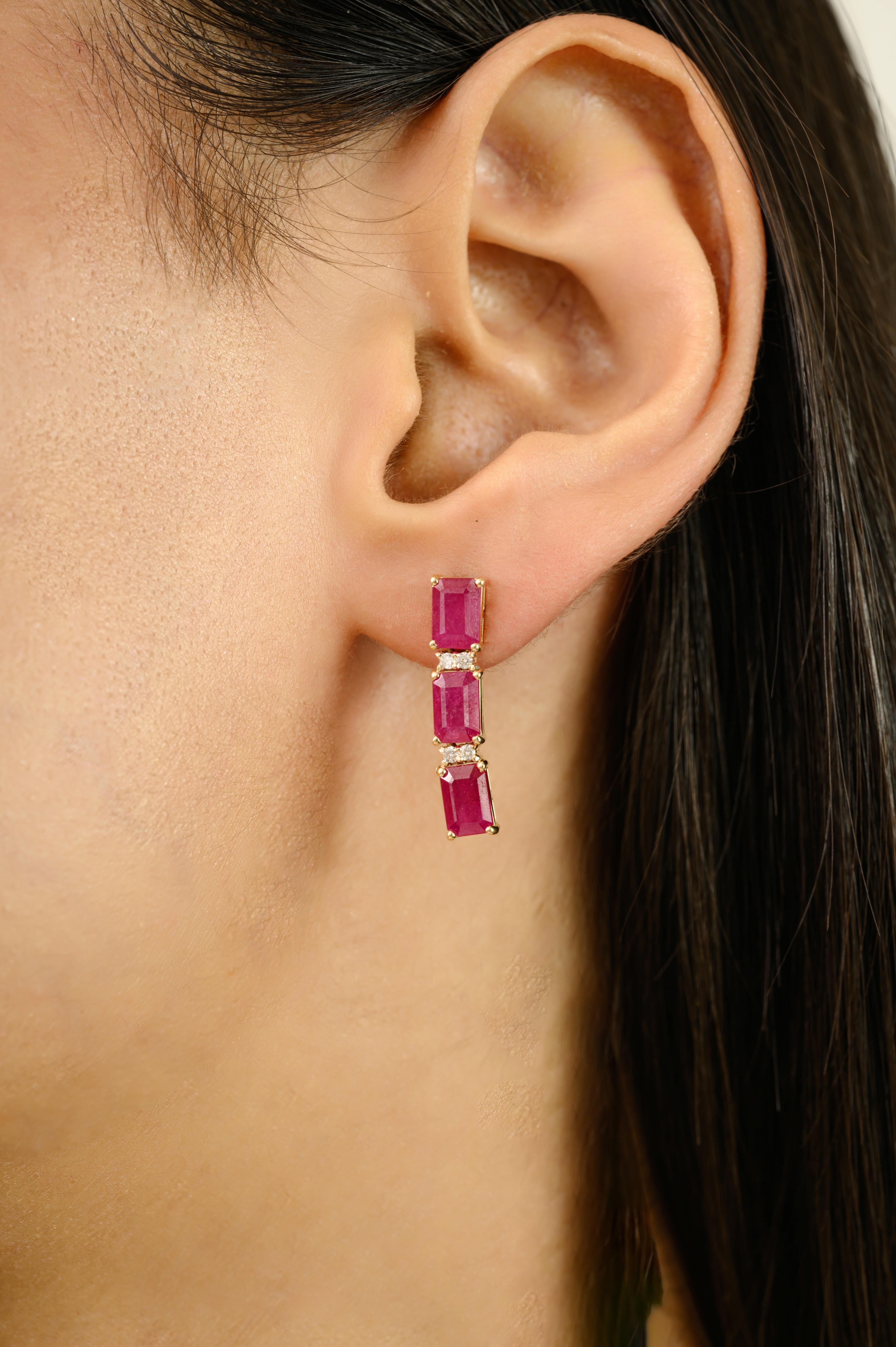 These 4.57Ct 14k Yellow Gold Natural Emerald Cut Ruby & Diamond Drop Earrings feature a modern bar design adorned with 4.57 carats of vibrant natural rubies and 0.09 carats of sparkling diamonds. Each earring showcases three beautifully cut rubies