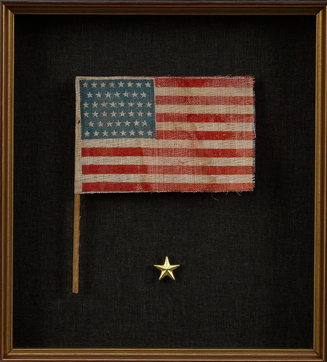 Presented is an American flag waver with 46 stars. The printed flag features a row pattern of stars against a dark blue canton, with thirteen red and white stripes completing the design. The small flag waver was a popular item in the early 20th
