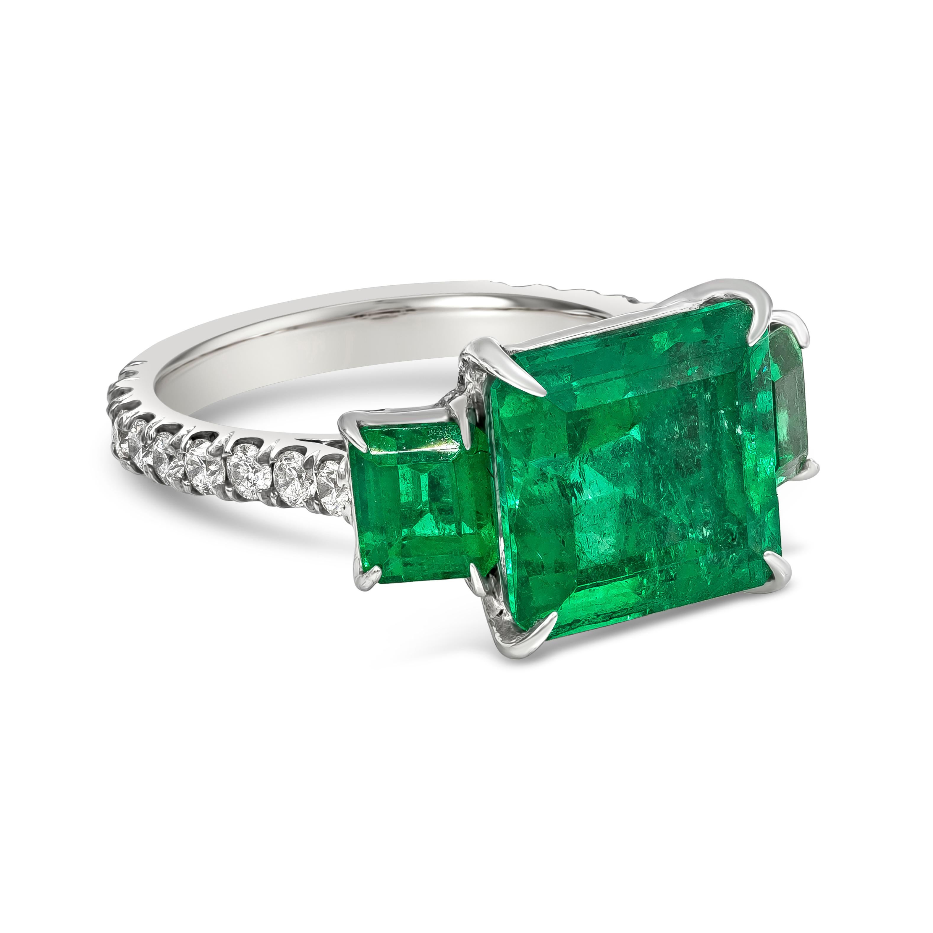 A color-rich and elegant three-stone engagement ring showcasing a GIA certified 4.82 carats emerald cut Colombian green emerald, set in a four prong basket setting. Flanked by two smaller emerald cut green emeralds on each side weighing 1.22 carats