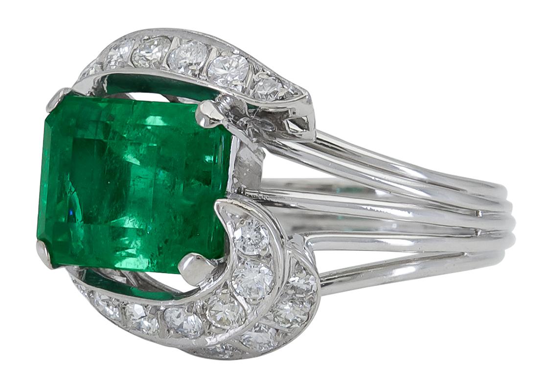 A fashionable cocktail ring showcasing a vibrant green emerald, surrounded in a ribbon-like design accented with brilliant diamonds. Set in a polished white gold mounting. 
Green emerald weighs 4.86 carats.
Size 8.5 US
Length: 0.68 inches