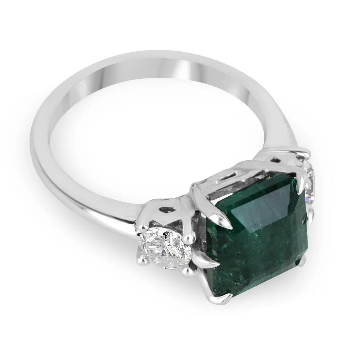 An intensified emerald and diamond three-stone ring. This exquisite piece features a large natural asscher cut emerald, that showcases a ravishing, deep green color with very good luster and clarity. Accenting the center stone are two large