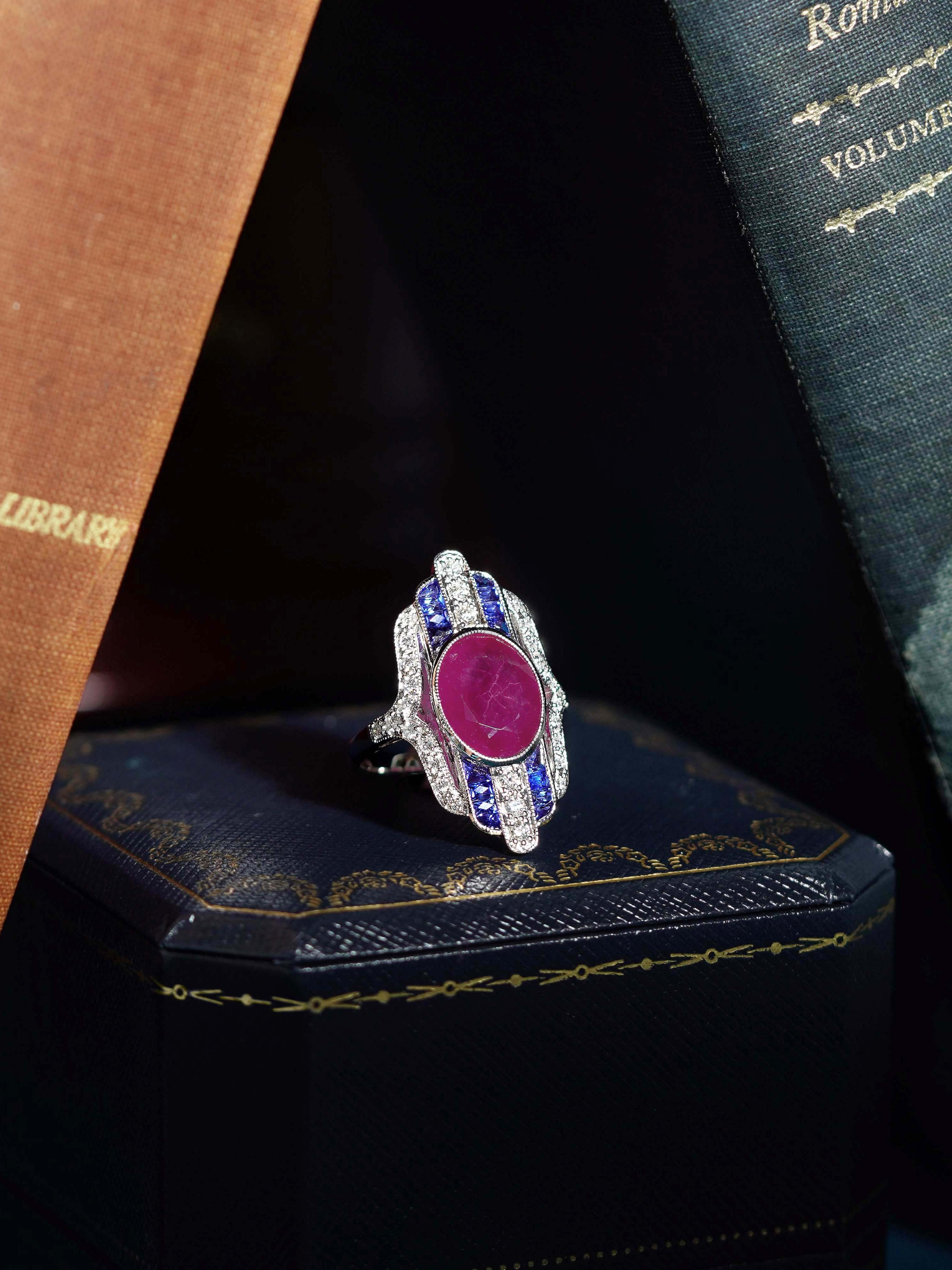 Bold and elegant, this Art Deco–style dinner ring features a rich, oval-cut ruby, certified and guaranteed for quality, bezel-set at the center. It is flanked by alternating French-cut sapphires and brilliant diamonds, arranged in an intricate,