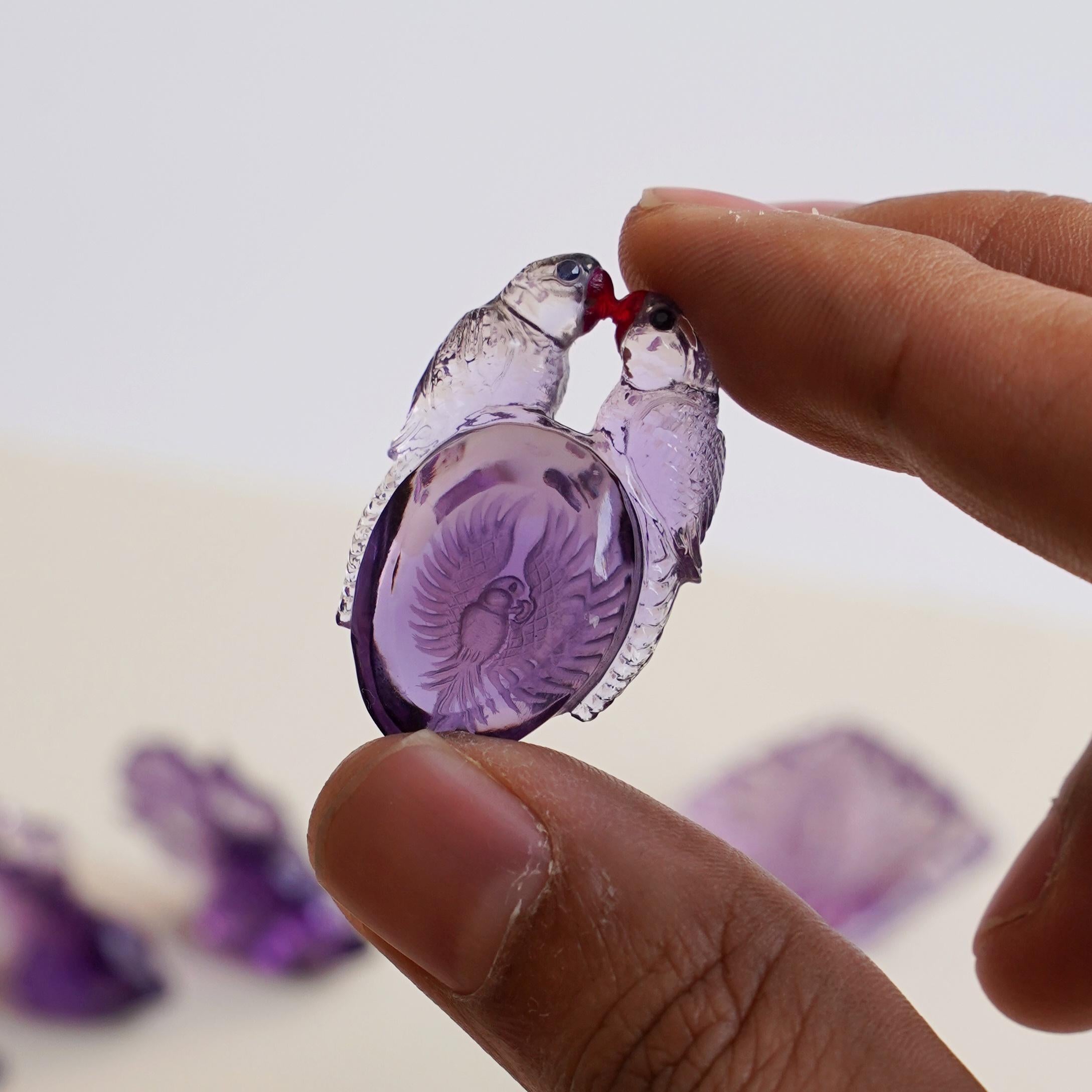 This love couple parrots pair carving on a natural Brazilian lavender amethyst is hand-carved by our expert lapidary artist in jaipur which transforms raw stones into unique art works. The baby parrot which is undercaved with reverse intaglio