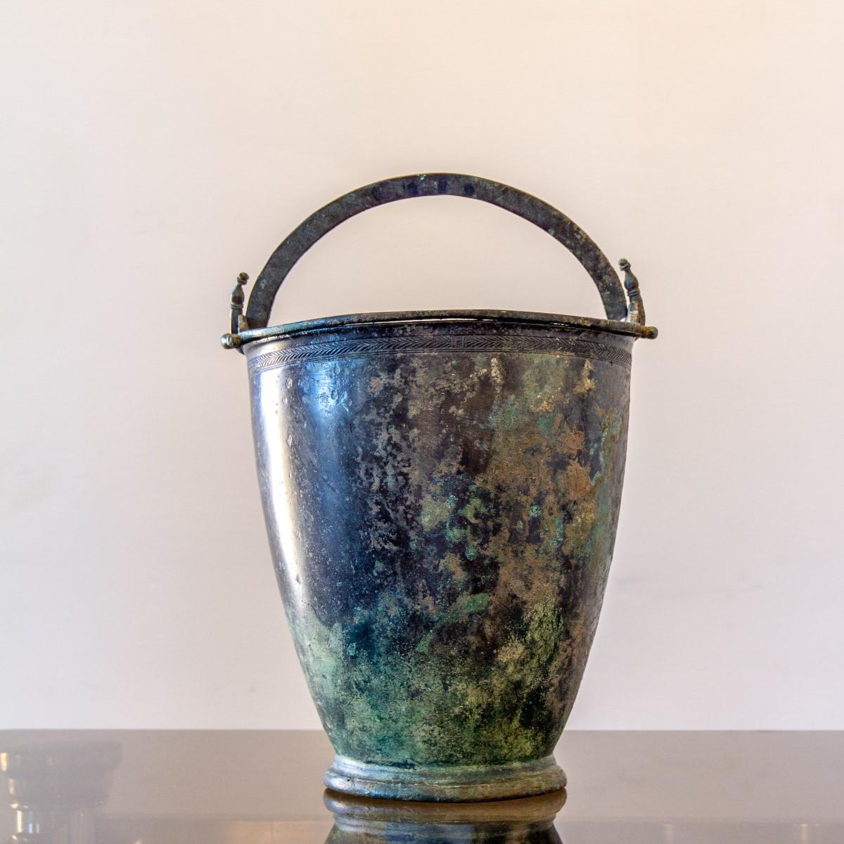 A 4th century BC Grecian bronze bucket with a circular foot and tapering cylindrical body. The feathered frieze around the top rim sits below two double rings with central carved palmettes and the two handles are omega shaped and hooked into loops,