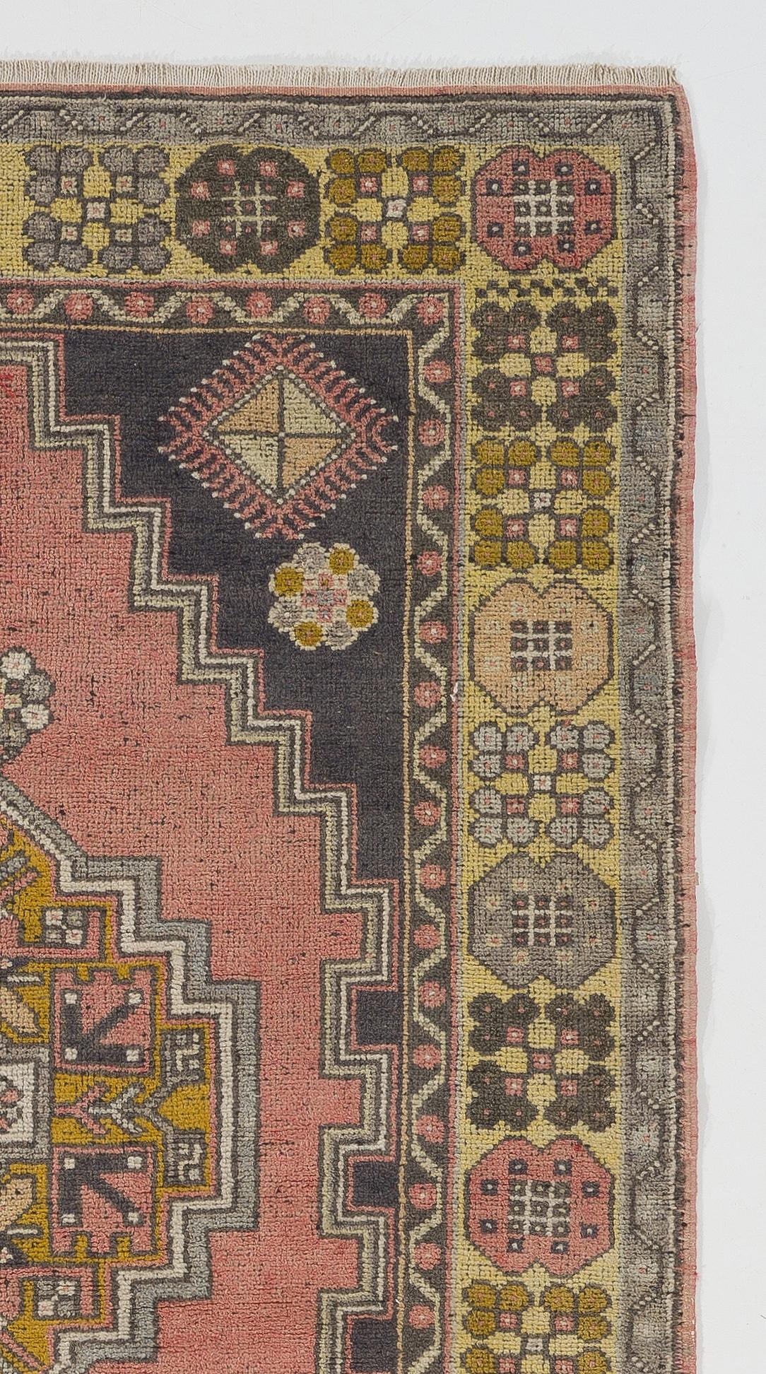 This vintage area rug was hand knotted in Turkey in the 1950s. It features a geometric medallion at the centre of two nested lozenge-shaped areas within a field in faded red and faded black colors featuring tassel-like motifs on their borders. The