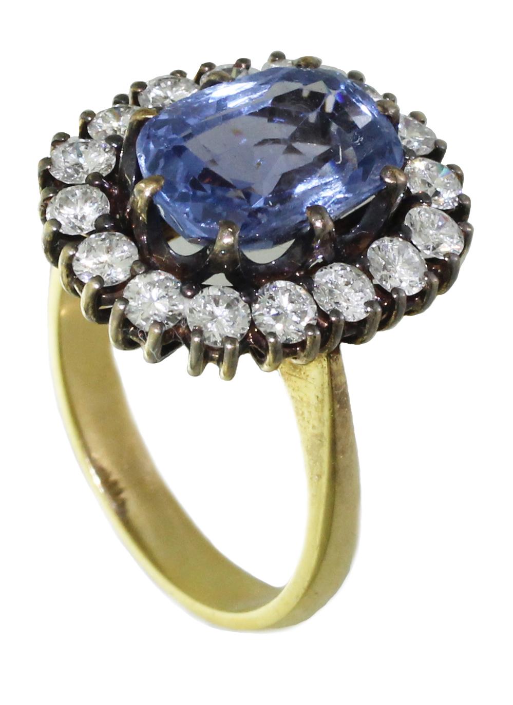 This precious cluster style ring features a 5 carat oval blue Ceylon Sapphire flanked by sixteen diamonds set in 18 yellow gold. Set to center with a single cushion shape old cut natural un enhanced Ceylon sapphire in an open back claw setting with