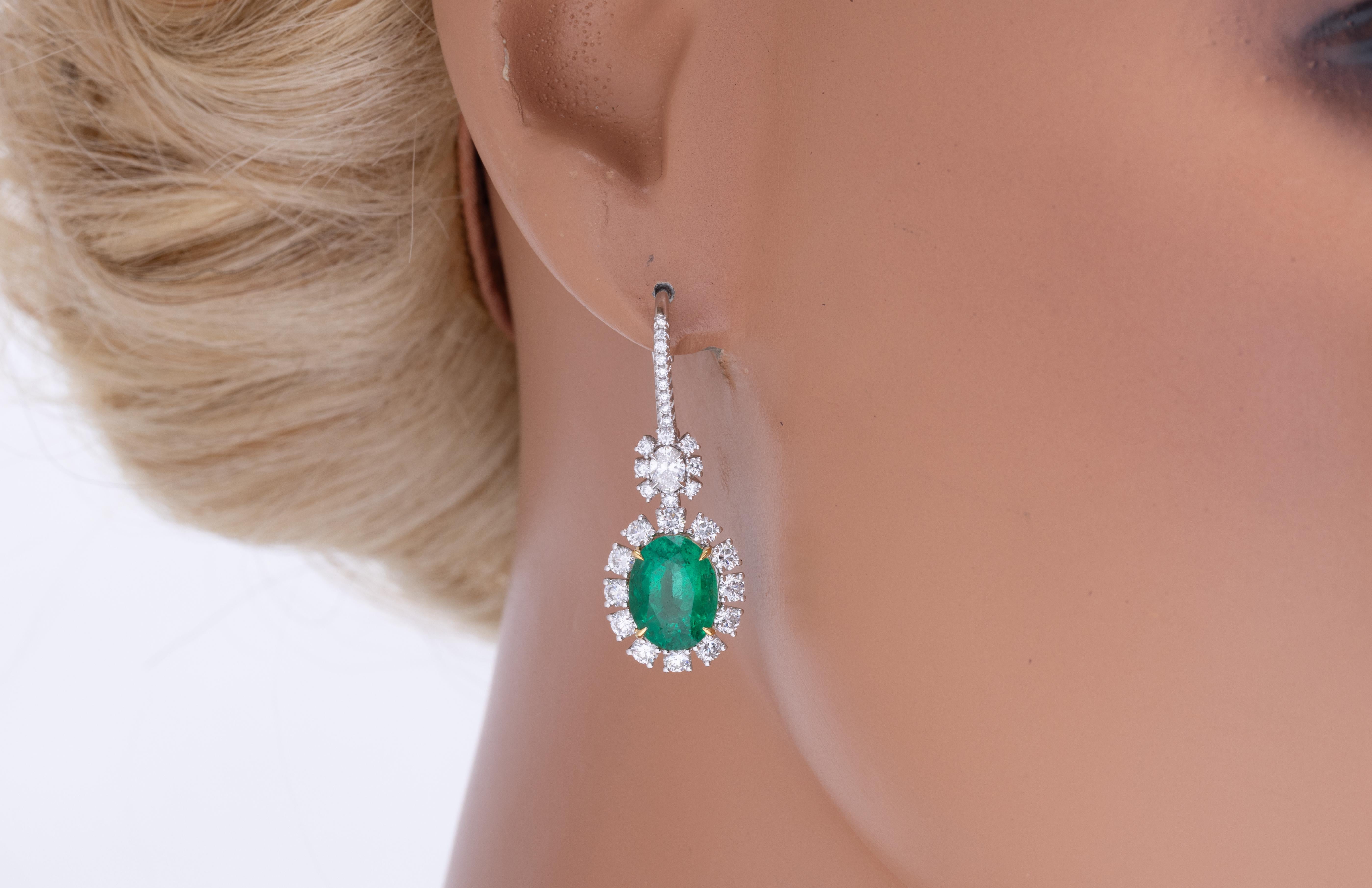 Questi squisiti orecchini presentano smeraldi ovali verdi dall'allure lussureggiante. Gli smeraldi sono completati da una scintillante aureola di diamanti naturali Whiting. Questi orecchini sono sapientemente realizzati in platino, con un cestino in