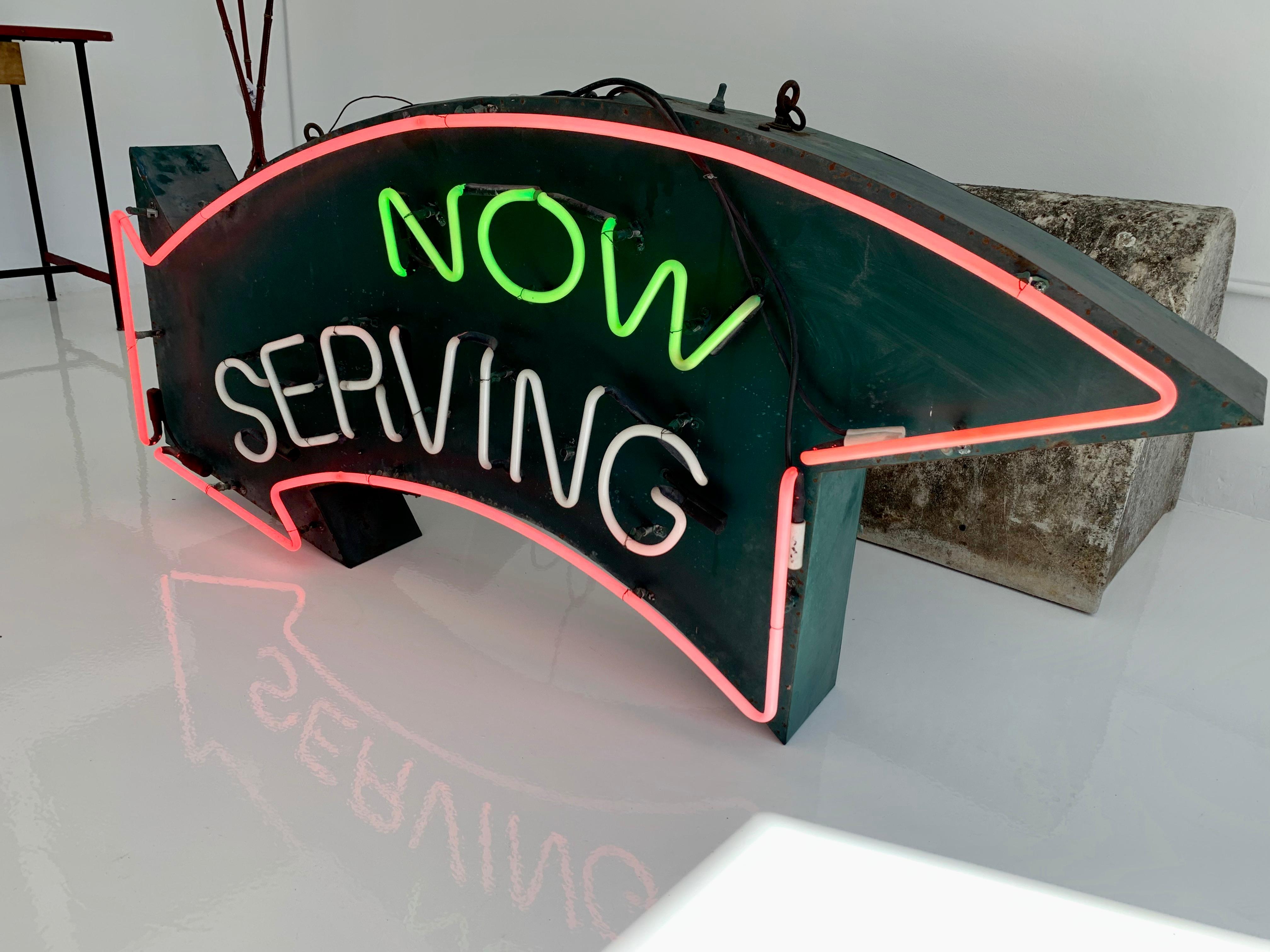 5 foot wide neon sign that says NOW SERVING. Vintage green metal arrow sign with neon lights attached. Hot pink neon borders the arrow as well as the word SERVING. The word NOW is in green neon. Able to turn off the wording so just the arrow lights