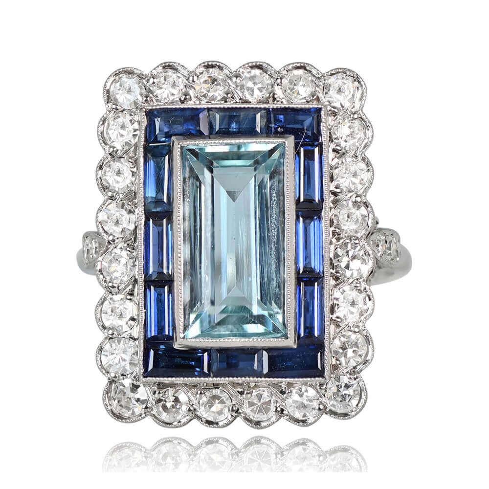 An emerald-cut aquamarine weighing around 5.00 carats takes center stage in this ring. The aquamarine is bezel-set and encircled by a double halo of baguette-cut natural sapphires and single-cut diamonds. A diamond-set fleur-de-lis motif graces the