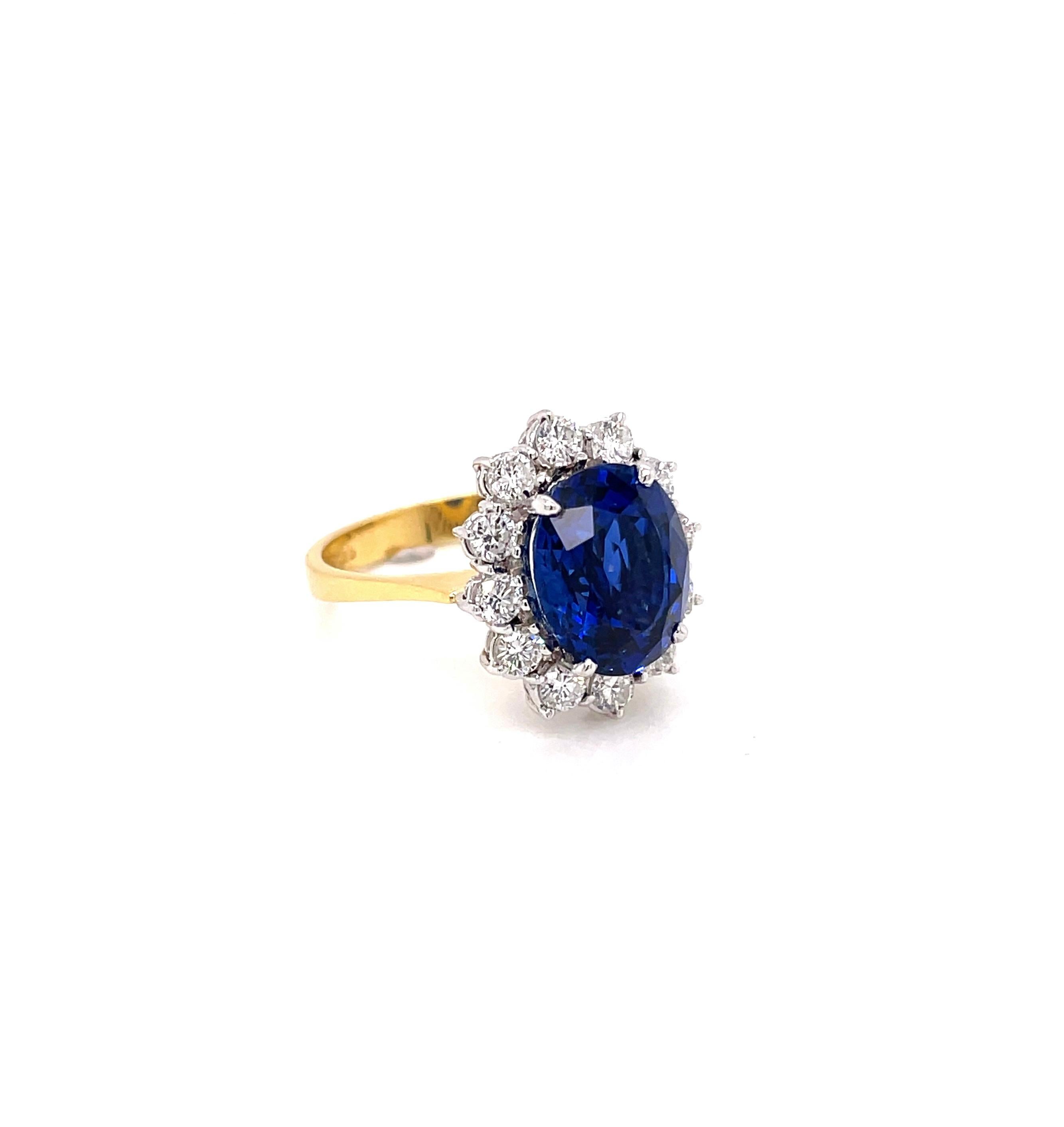 Princess Diana inspired cluster engagement ring set with an exquisite oval shaped royal blue sapphire weighing 5.03 carats in a four claw, open back setting. The gorgeous stone is beautifully surrounded by twelve fine quality round brilliant cut
