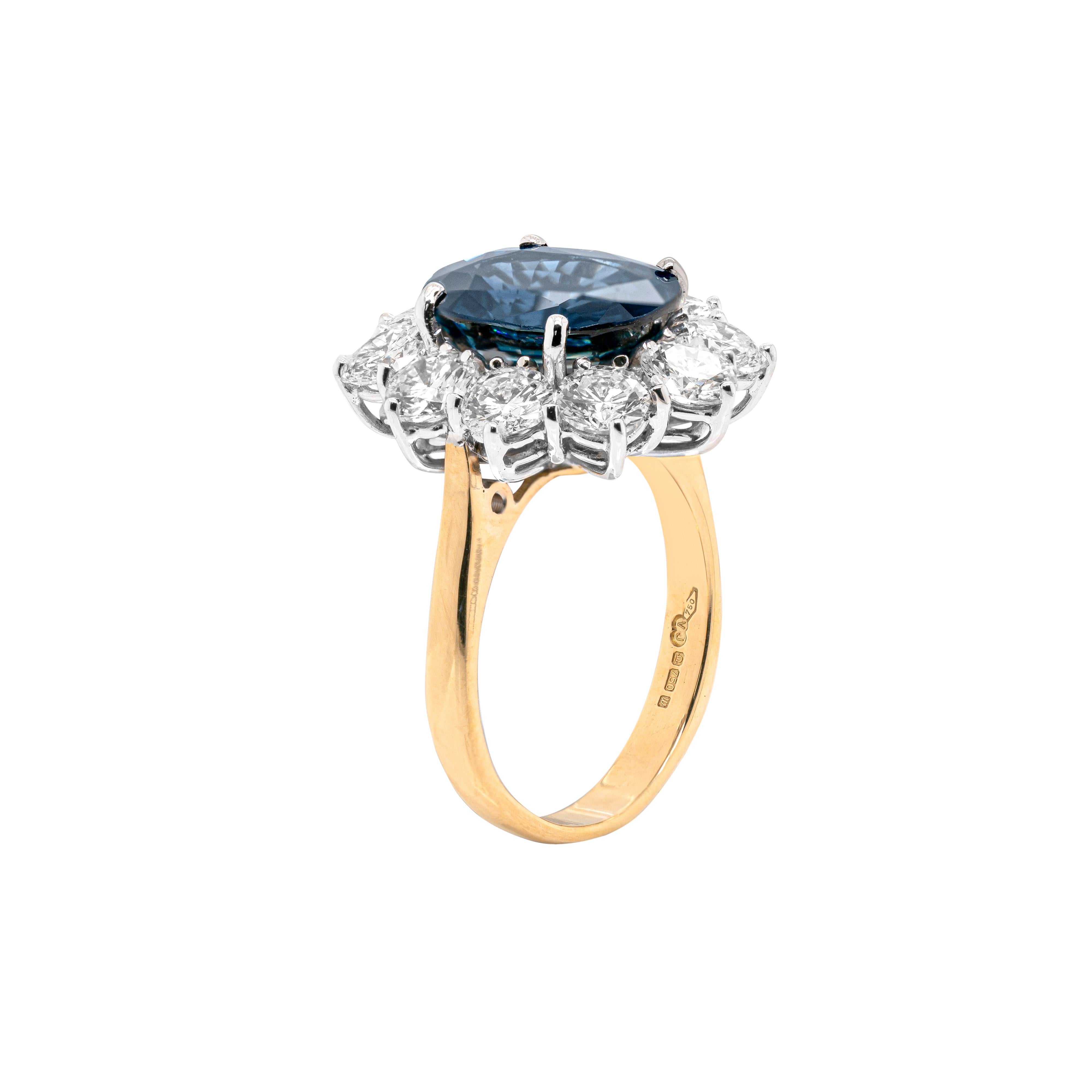 This incredible engagement cluster ring features a wonderful oval shaped blue sapphire weighing 5.03 carats, in a four claw, open back setting. The impressive stone is beautifully surrounded by ten round brilliant cut diamonds weighing a total