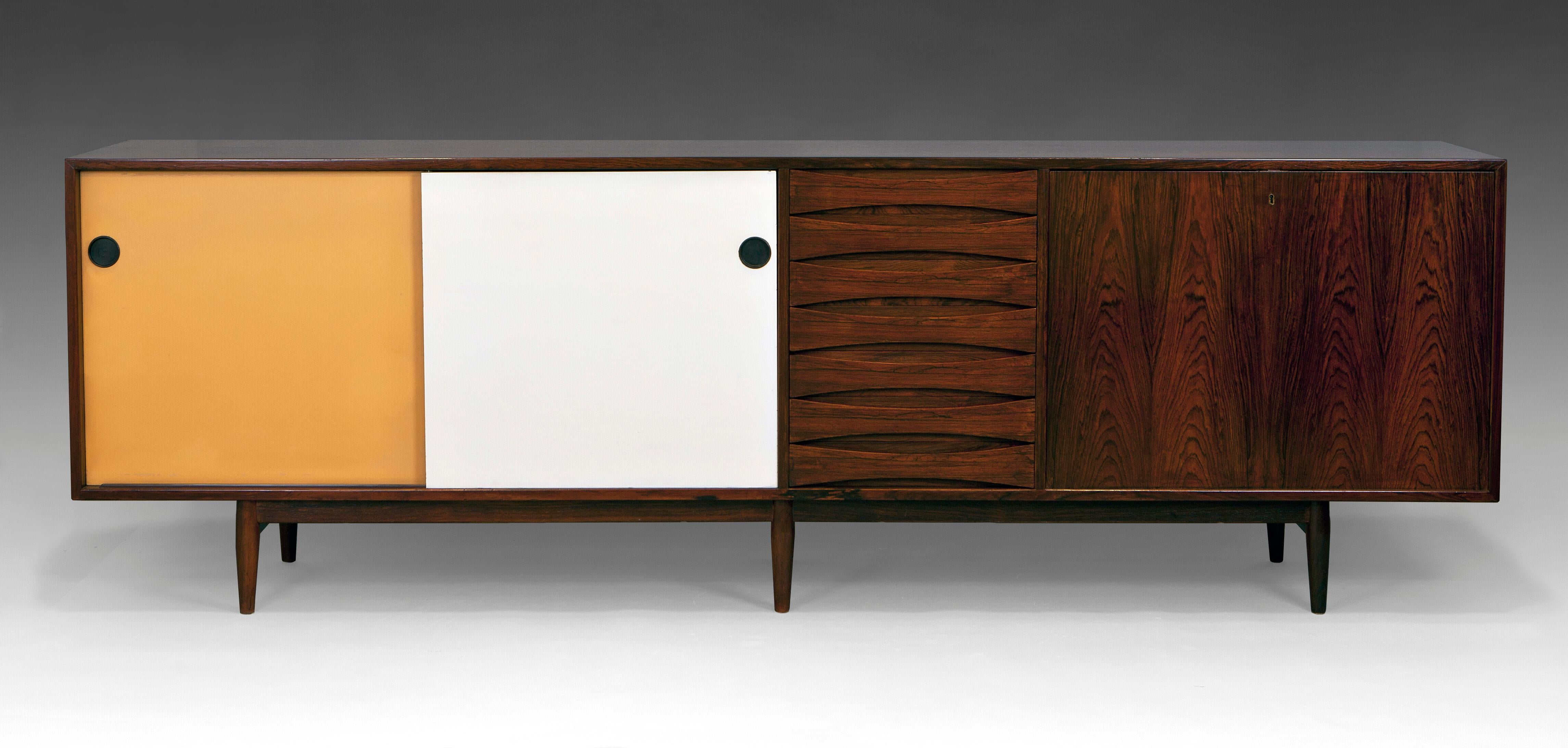 Rosewood “29 A” Sideboard by Arne Vodder for Sibast. Denmark, late 1950s. Freestanding rosewood sideboard on six legs. Front with two sliding doors painted in white and ocher. Central section with seven drawers with curved handles. Bar section with