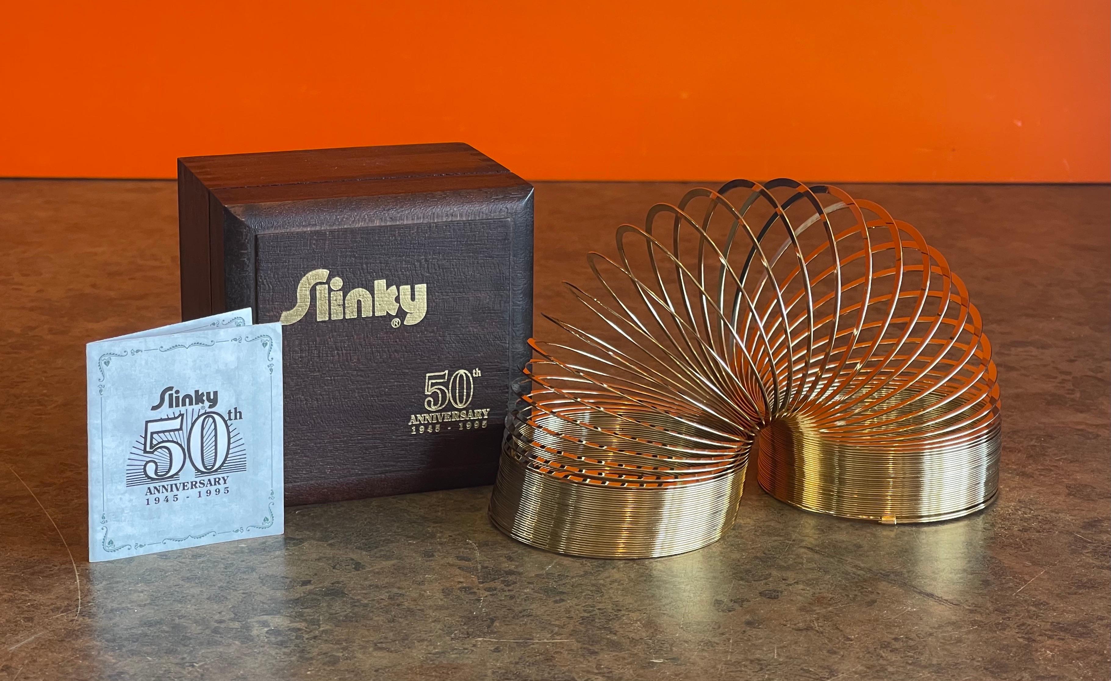Like new 50th Anniversary gold-plated brass Slinky toy in wood box, circa 1995. This item is in original plastic packaging an has never been played with; the piece comes with the original brochure / song sheet. The Slinky was released in 1995 to