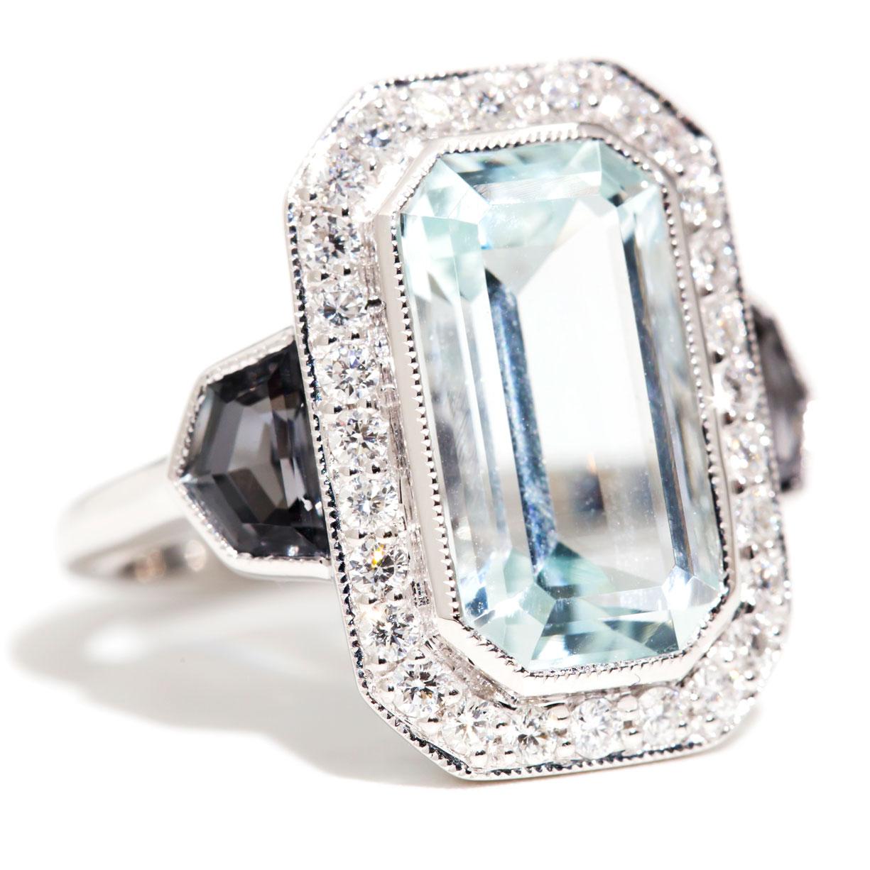 This wondrous art deco inspired ring is forged in 18 carat white gold and features a  flawless, bright light blue 5.10 carat emerald cut Aquamarine. The captivating Aquamarine is encompassed by a border of sparkling round brilliant cut diamonds, and