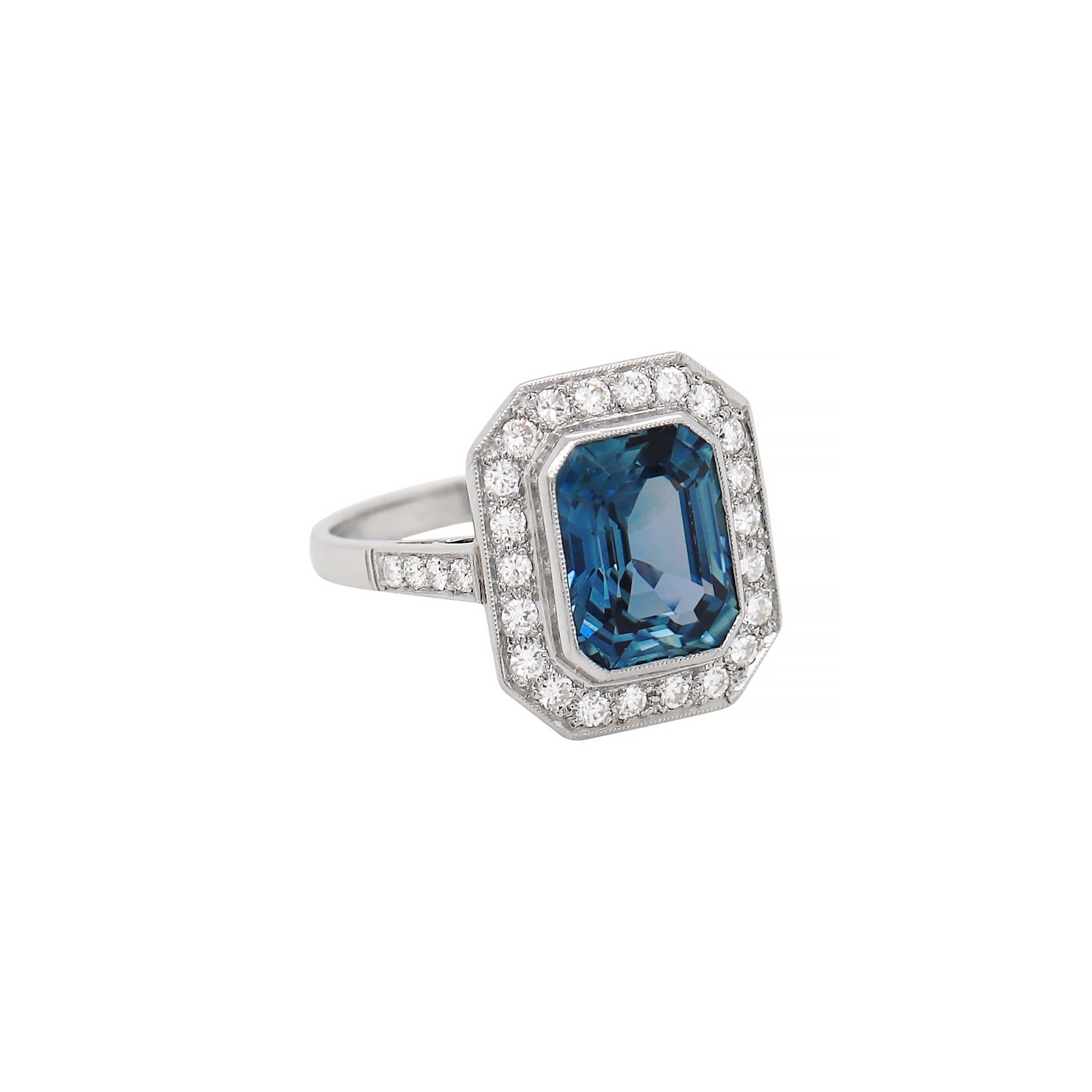 This exquisite ring features a beautiful transparent blue natural unheated emerald cut sapphire weighing 5.12ct in a rub over, open back setting. The incredible stone is surrounded by 22 fine quality round brilliant cut diamonds, all grain set in