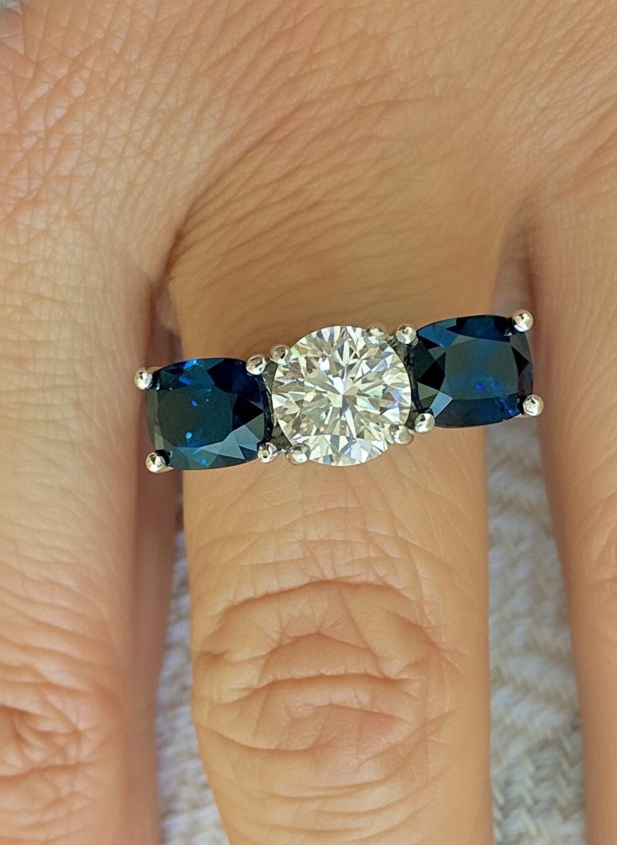 Cette magnifique bague à trois pierres en or blanc 18 carats est ornée d'un diamant rond de taille brillant de 1,73 carats (pureté améliorée), au centre, ainsi que de deux magnifiques saphirs naturels bleu royal de taille coussin pesant au total