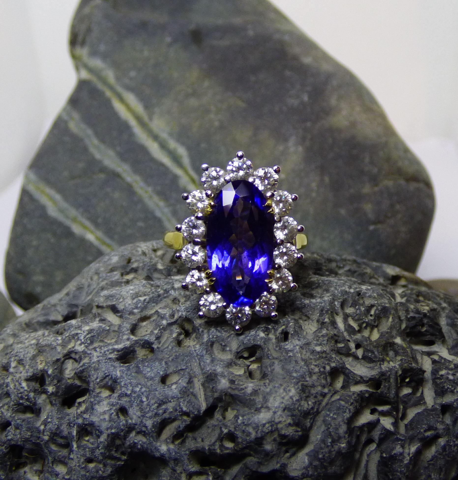 A bright, clean, colourful Tanzanite weighing 5.20ct. is surrounded by 14 Diamonds with a total weight of 1.40ct. The total surface front of the ring is  20X13mm. It is handmade in 18ct. yellow white gold.