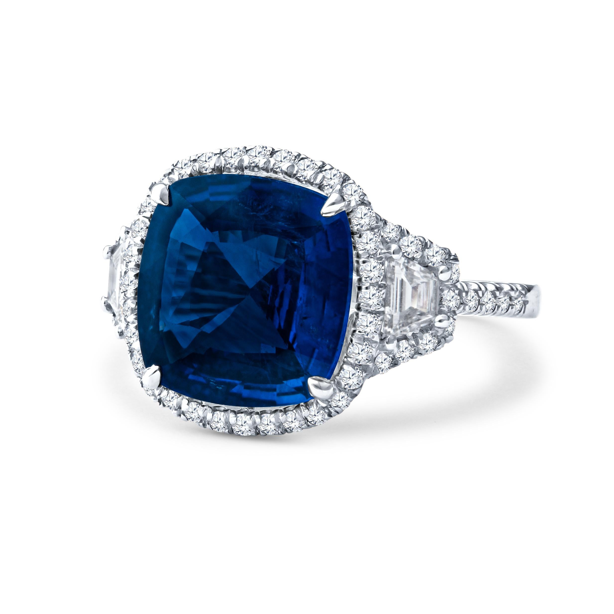 This exquisite ring features a natural 5.21ct blue sapphire center stone in a cushion shape, bordered by two step cut trapezoid diamonds with a total weight of 0.29ct, and 0.51ct total weight in round accent diamonds forming the halo around the